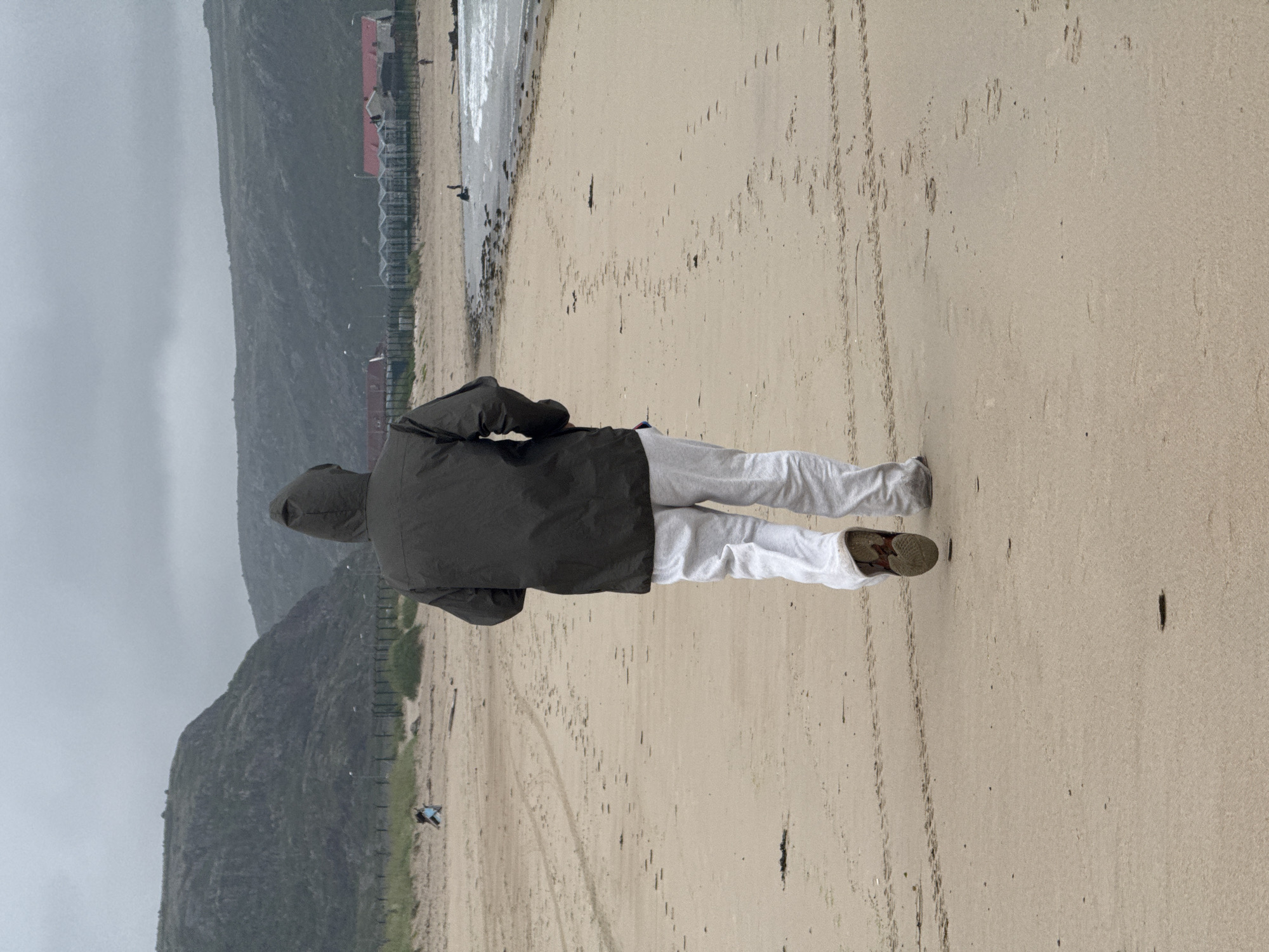 A person in a dark hooded jacket and light-colored pants walks away from the camera on a sandy beach. The beach has a rough texture with scattered debris and numerous footprints. In the background, a coastline with rocky hills rises under a cloudy, overcast sky. To the right, the ocean meets the shore with moderate waves. Further in the distance, a line of buildings with red roofs and glass-paneled structures is visible along the coast, suggesting a populated area within the city of Teriberka, Russia. A fence runs along the edge of the beach, separating it from the built-up area. Several other figures can be seen as small shapes on the beach and near the water. The overall atmosphere is somber and possibly cool, indicated by the clothing and the weather.