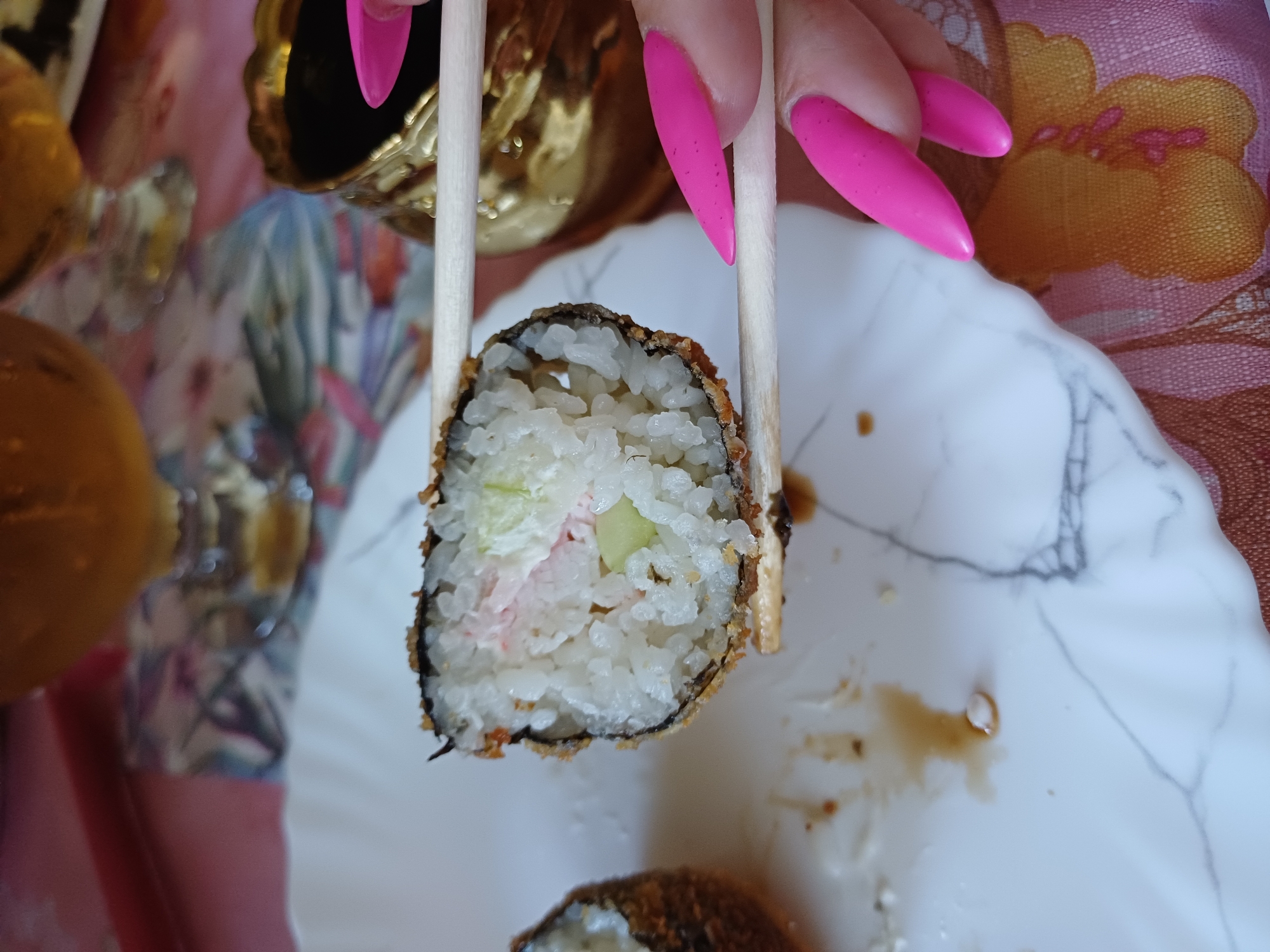 In an indoor setting, a close-up shot captures a piece of sushi being held by chopsticks over a white, marble-patterned plate. The sushi roll is tempura-fried, with visible rice and fillings of pink and green. To the right, a person's hand with bright pink, stiletto-shaped fingernails is visible, with the chopsticks held between the index and middle fingers. In the background, a golden container and glassware are partially visible. The surface beneath the plate has a patterned fabric in pink and yellow. The scene appears to be a dining setting, possibly a restaurant or home.