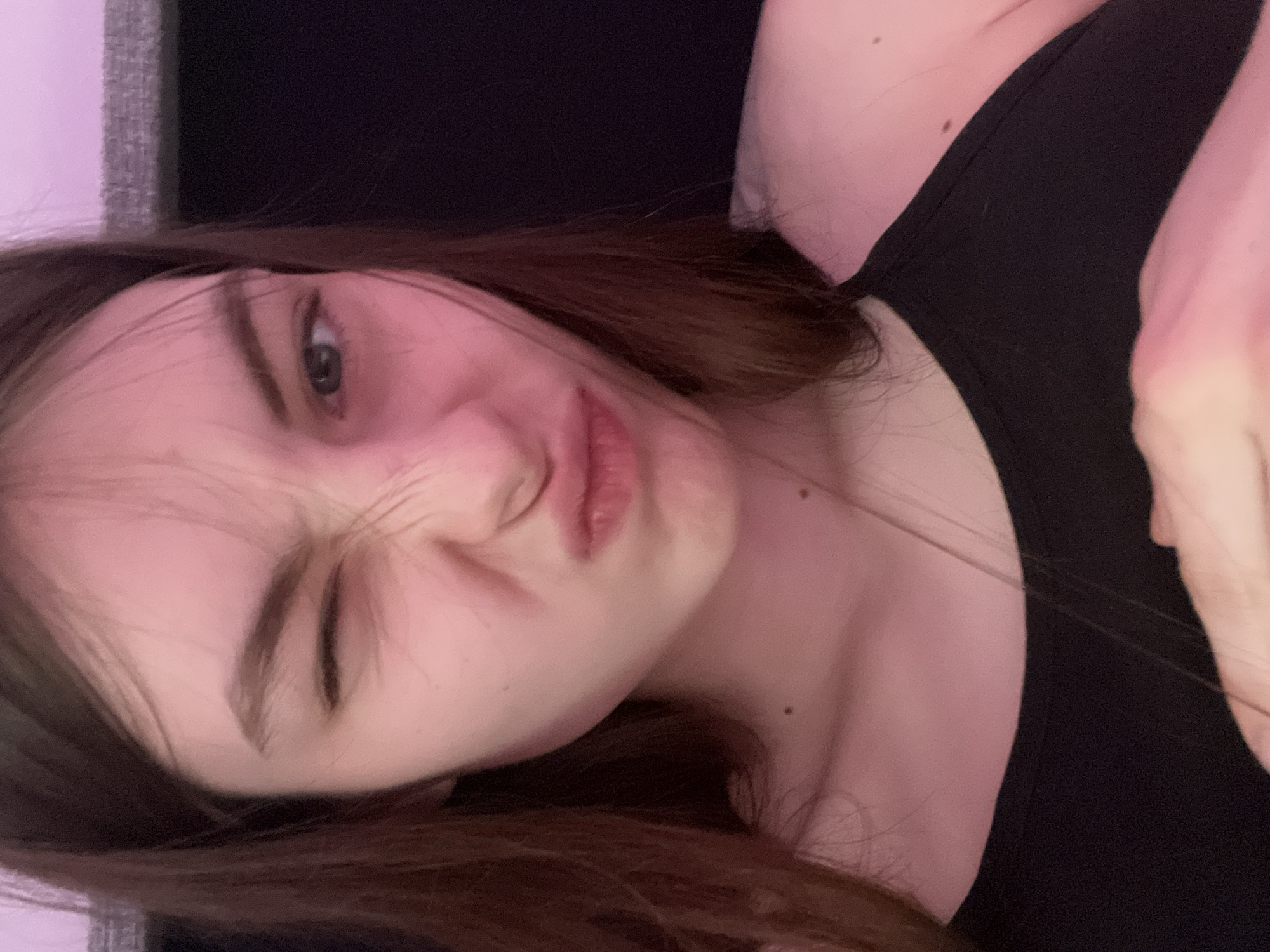 A close-up selfie depicts a young, light-skinned woman with brown hair. She is wearing a black tank top. Her face displays a playful expression, with one eye squinted shut and her lips pursed in a pouting manner. The image is slightly tilted. The background is dark and indistinct. There is no information to suggest the picture was taken in Saint Petersburg, Russia.