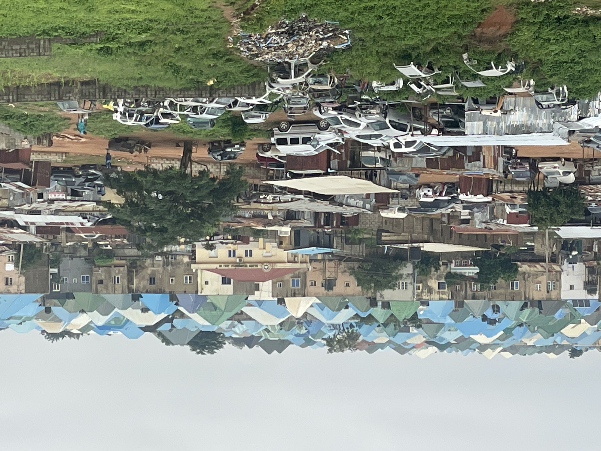The image depicts a densely populated urban landscape in Abuja, Nigeria, characterized by a multitude of residential buildings with distinctively colored, often conical, roofs in shades of blue and green. The architecture suggests a mix of modern and older structures, with some buildings appearing more developed than others. In the foreground and mid-ground, a significant number of abandoned and dismantled vehicles are visible, scattered across grassy areas and what appears to be a makeshift junkyard or scrap area. Several of these vehicles are in various states of disrepair, with missing parts and exposed interiors. The environment is overgrown with vegetation, indicating a possible lack of maintenance or a deliberate integration of greenery. A few individuals are present in the scene, some appearing to be working or moving around the vehicles, while others are more distant, interacting within the residential area. The overall impression is one of a bustling, possibly lower-income or developing section of the city, where discarded vehicles are a prominent feature of the landscape, contrasting with the more uniformly roofed residential structures.