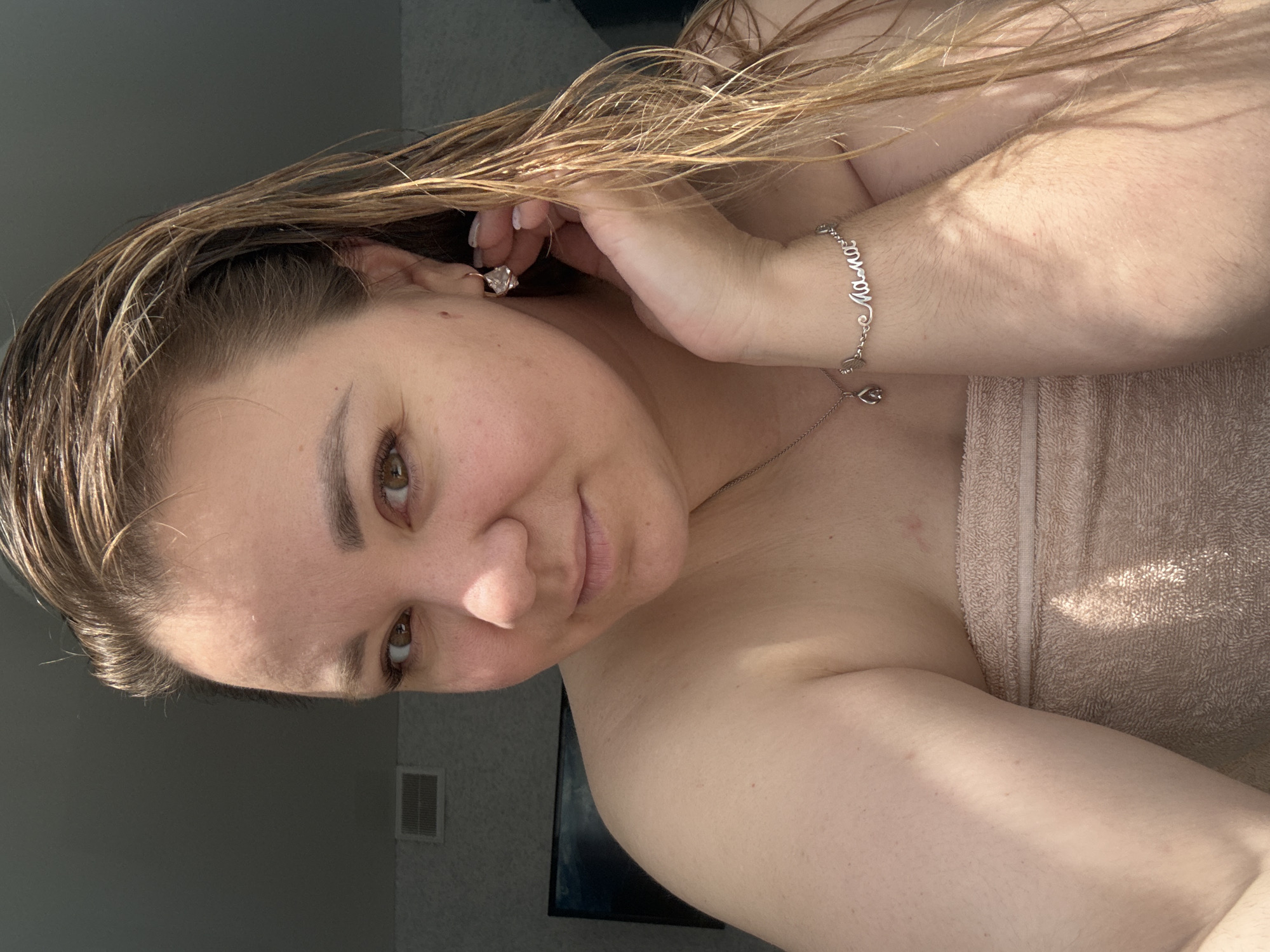 A woman with wet, brown hair is captured in a close-up, head-and-shoulders shot. She is looking towards the camera with a slight smile. She is wearing a light brown towel wrapped around her chest and has on jewelry, including a necklace with a teardrop pendant and a bracelet with the word "mama" in cursive. Her right hand is near her face, touching her ear where she wears a small, clear earring. The background appears to be an indoor setting, with a gray wall and a ventilation grate visible on the left side, and a dark television screen behind her on the left. Sunlight is casting shadows across her shoulder and arm. The image is a selfie, taken in a personal, domestic environment.