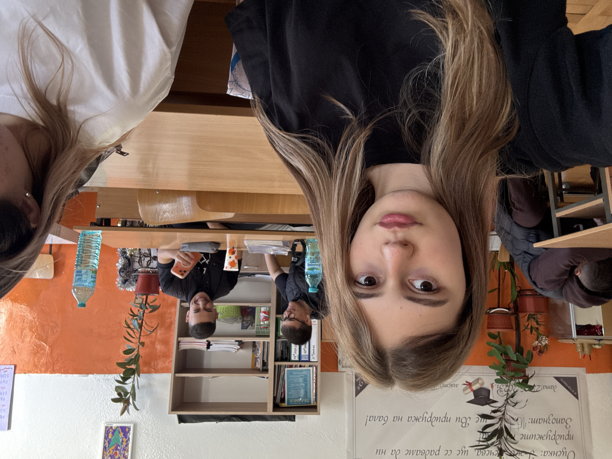 The image depicts three young adults in what appears to be a classroom in Panagyurishte, Bulgaria. A young woman with long, light brown hair is positioned in the foreground. To her left, another young woman is partially visible. In the background, a young man is visible, looking at his phone and smiling. Another young man is visible behind a desk with books in the background. The setting features a desk with books and a water bottle, a bookshelf filled with various books, a plant, and an orange wall. A poster with Bulgarian Cyrillic writing and a graduation cap illustration is visible in the foreground.