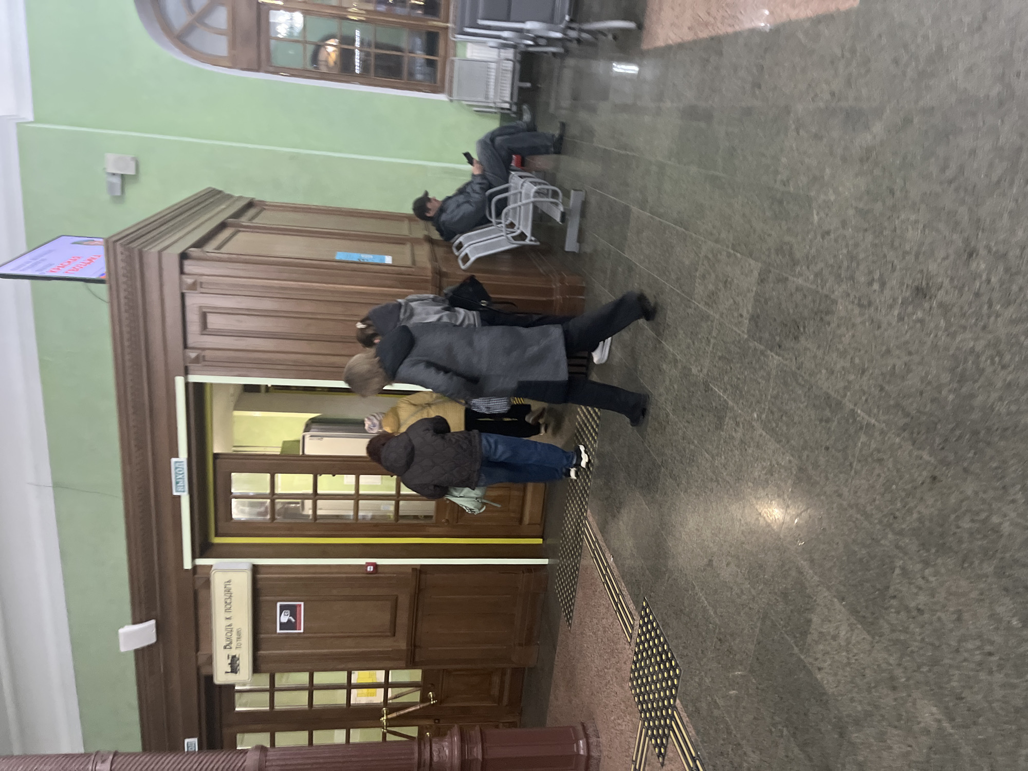 The image depicts the interior of Perm-2 railway station in Perm, Russia. Several people are present, some standing near a wooden doorway with a sign written in Russian reading "Exit to trains". One person sits on a metal bench, looking at a mobile device. The interior has light green walls, brown wooden trim, and a gray tiled floor. Some seating is arranged near a window in the upper part of the frame.
