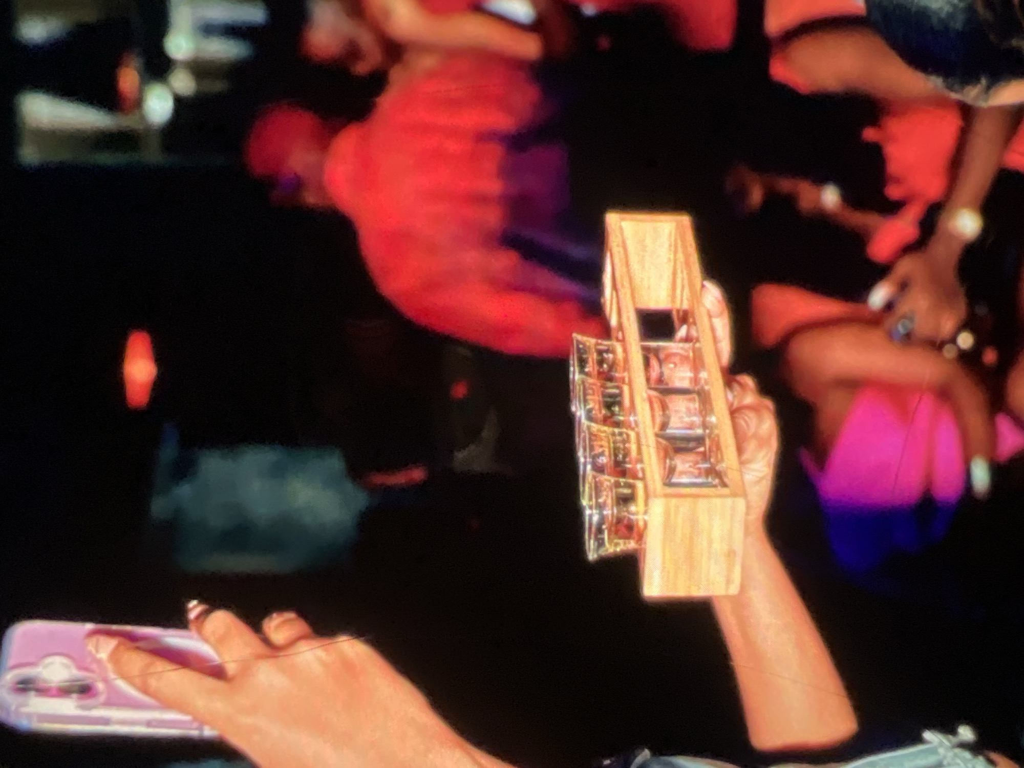 The image depicts a dimly lit scene, likely an indoor venue, possibly a club or bar, in Lagos, Nigeria. In the foreground, a hand holding a pink smartphone is visible, its screen reflecting a purple case. This hand is positioned to the left of a wooden tray holding several shot glasses filled with amber liquid, suggesting a celebration or social gathering where drinks are being served. The background is blurred but shows several individuals. One person is prominently visible in a bright red top, facing away from the camera, with another person partially visible in a vibrant pink top. The overall impression is one of a lively social event, captured in low light conditions.