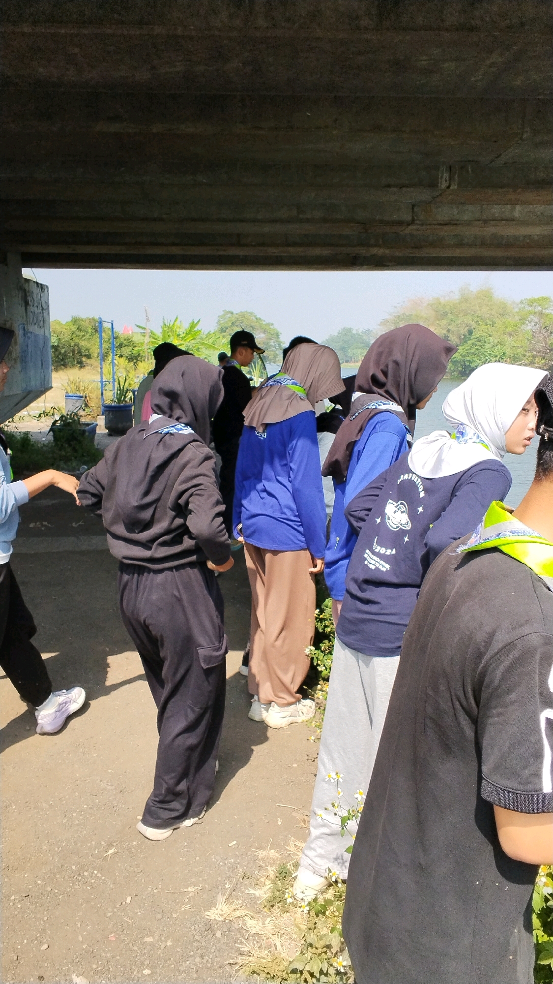 The image depicts a group of people, likely students based on their attire, gathered under a bridge. The setting appears to be outdoors, with vegetation and a body of water visible in the background. The ground is unpaved, with dirt and gravel. Several individuals are wearing headscarves and loose-fitting clothing. One person in the foreground is wearing a black t-shirt and another person to their left is wearing a black hooded jacket and pants. The overall atmosphere suggests a casual gathering or activity.