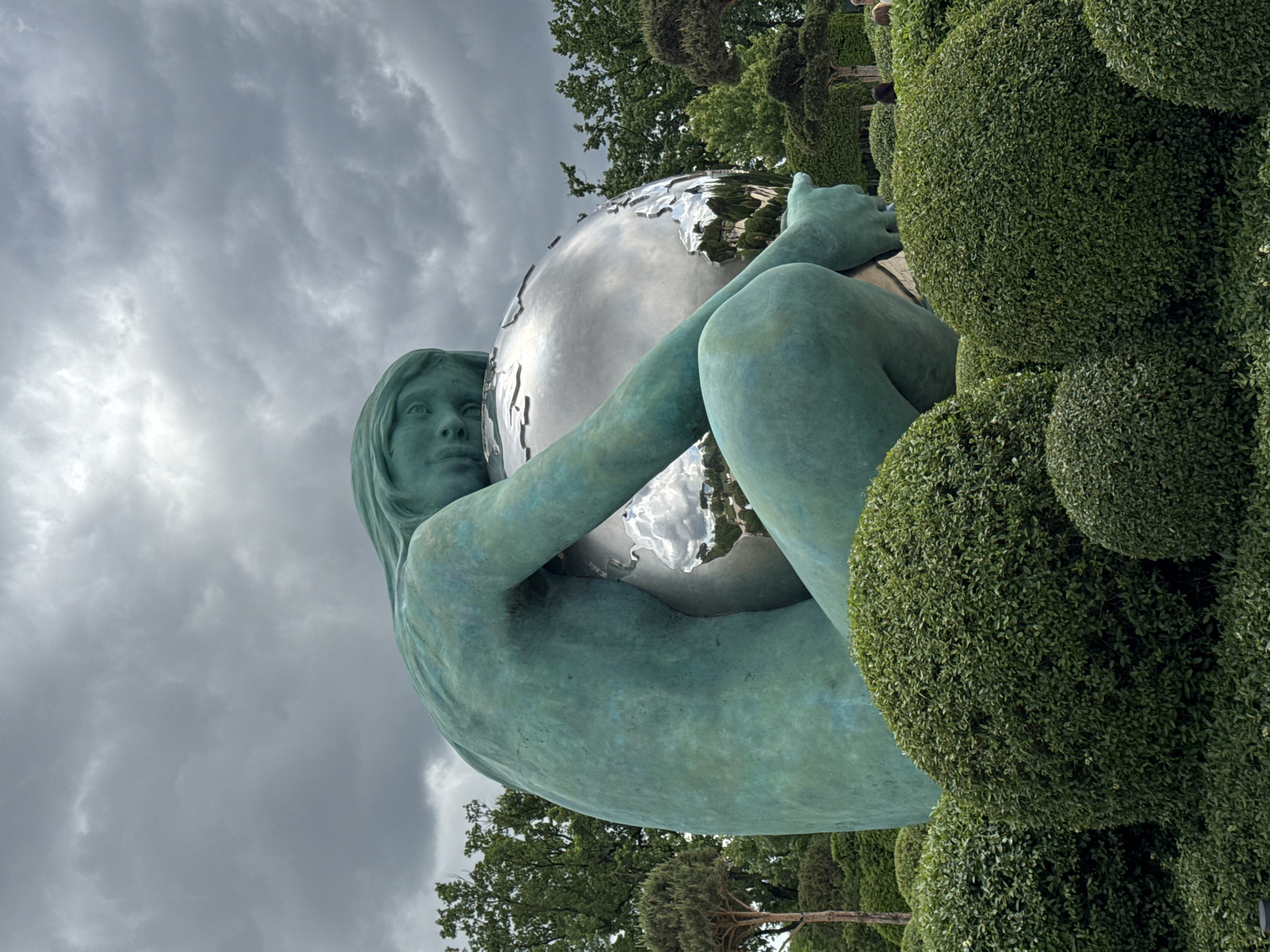 A low-angle, eye-level shot shows a statue of a woman holding a globe, located in Krasnodar, Russia. The statue of the woman is a weathered, green-blue color. She is seated and holds a silver, reflective globe that shows images of the park behind her. She looks upward to the left. Behind and to the right of the statue is a green bush in the shape of several round balls stacked on top of each other. The sky is overcast and gray. Trees are visible in the background.