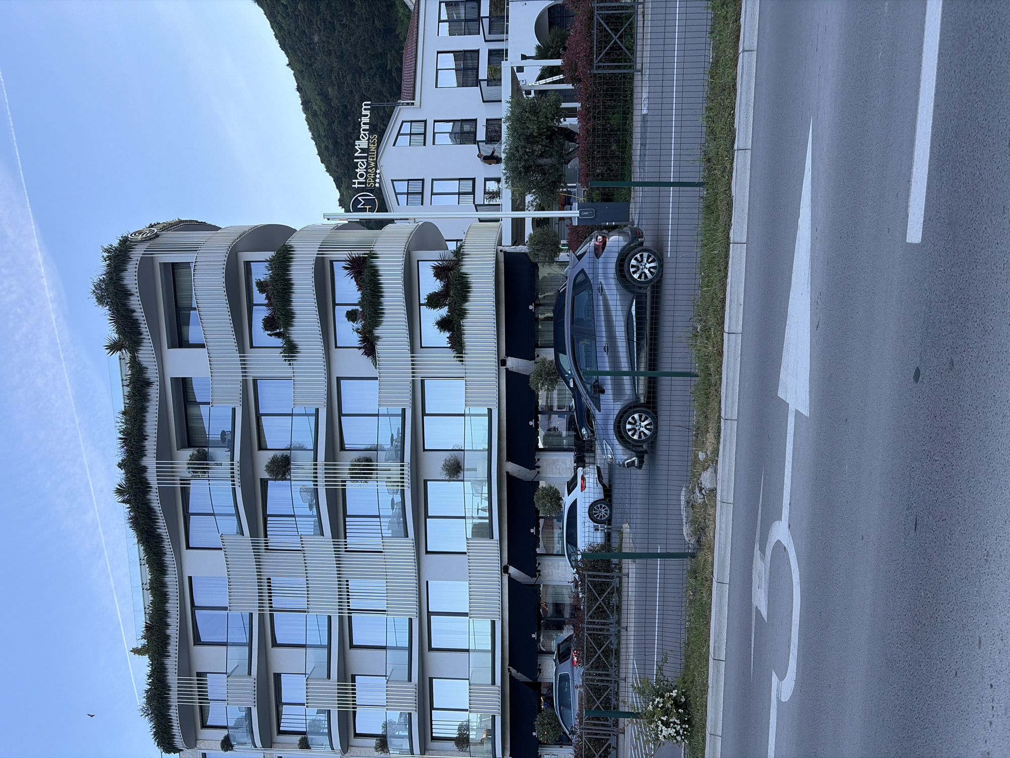 The image captures a street scene in Budva, Montenegro, featuring the Hotel Millennium. The hotel is a modern, multi-story building with curved balconies decorated with greenery. Several cars are parked along the sidewalk in front of the hotel. The road has white directional arrows painted on the asphalt. The sky is blue and clear.