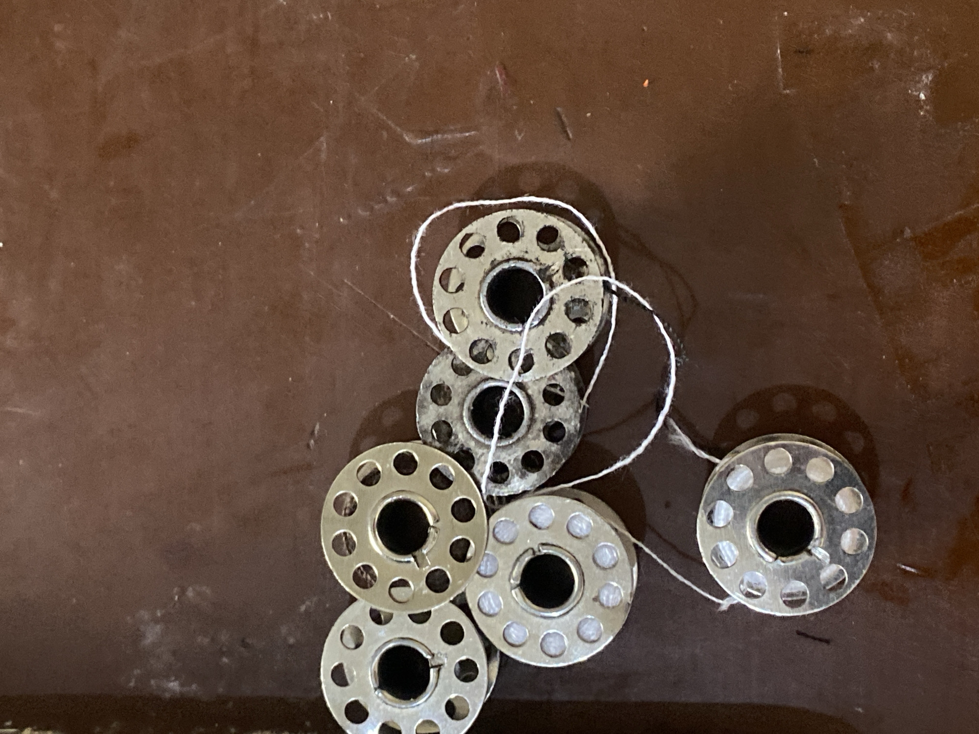 A close-up, overhead shot displays a collection of metallic sewing machine bobbins scattered on a dark, reflective surface. Six bobbins are visible, some with white thread wound around them. The bobbins are circular with multiple holes and a central opening. The background surface appears to be a polished wood or laminate with some scuff marks and dust. The lighting is uneven, casting shadows and creating highlights on the metallic surfaces. The image is taken from an elevated perspective, focusing on the arrangement of the bobbins. There is no indication of location within the city of Jauro Yinu, Nigeria.