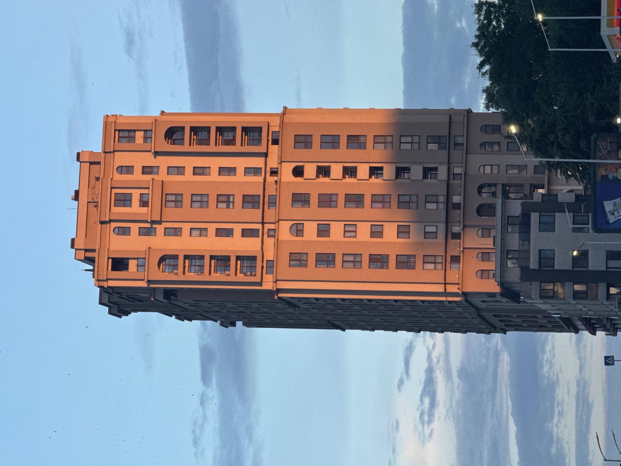 A tall, light-colored apartment building is prominently featured against a blue sky with scattered clouds. The building exhibits architectural elements such as arched windows and balconies, with a portion of its facade illuminated by the warm glow of sunset. The lower part of the image reveals a darker, modern building adjacent to the taller structure, along with glimpses of trees and streetlights. The scene is set in the city of Burgas, Bulgaria.