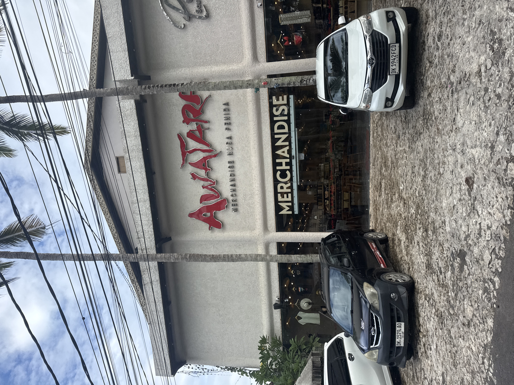 This outdoor image depicts a commercial building in Kampung Toyapakeh, Indonesia, hosting an "Awatar" merchandise shop. The building's facade features a large red "Awatar" sign above the word "Merchandise," with palm tree trunks framing the building's front. Two cars are parked in front of the shop: a white car with the license plate "DK 1942 FCD" and a black car with the license plate "DK.1579. AO." Power lines and palm fronds obscure part of the sky above the building. The ground appears to be paved with stone. The image is oriented vertically.