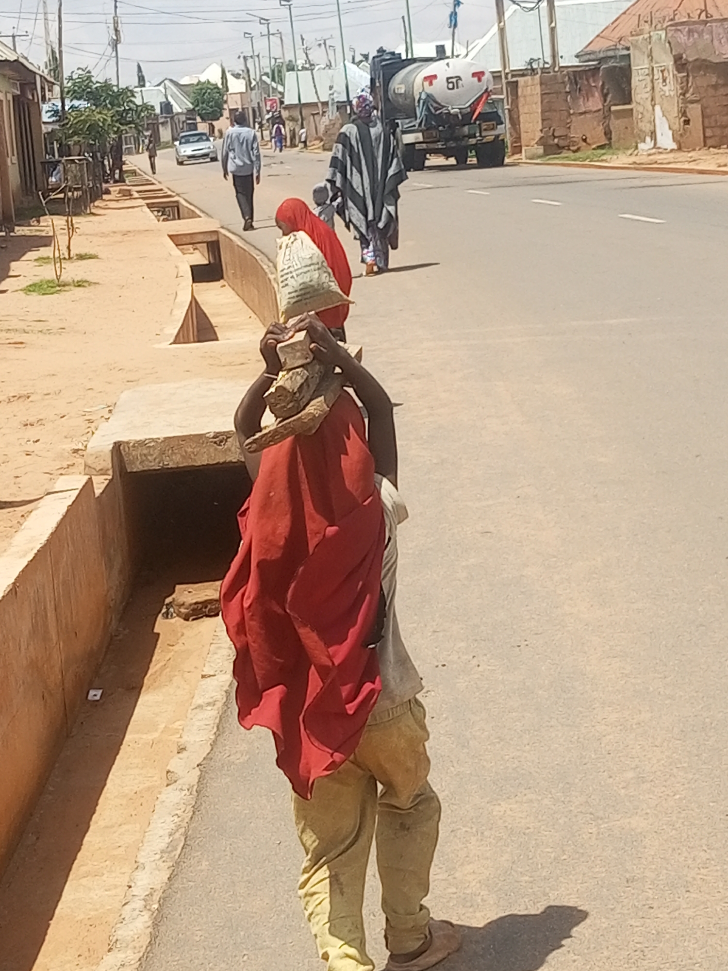 In the city of Gombe, Nigeria, a young person is walking down a paved road carrying a bundle of wood on their head. The person is wearing a red garment draped over their shoulders and a light-colored shirt underneath, along with loose-fitting tan pants. They are walking away from the camera towards a white truck with "5T" written on its side, which is parked on the right side of the road. In the background, there are buildings and other people walking along the road. The scene appears to be during the daytime, with clear weather. A drainage ditch runs along the left side of the road, with some vegetation growing in and around it.