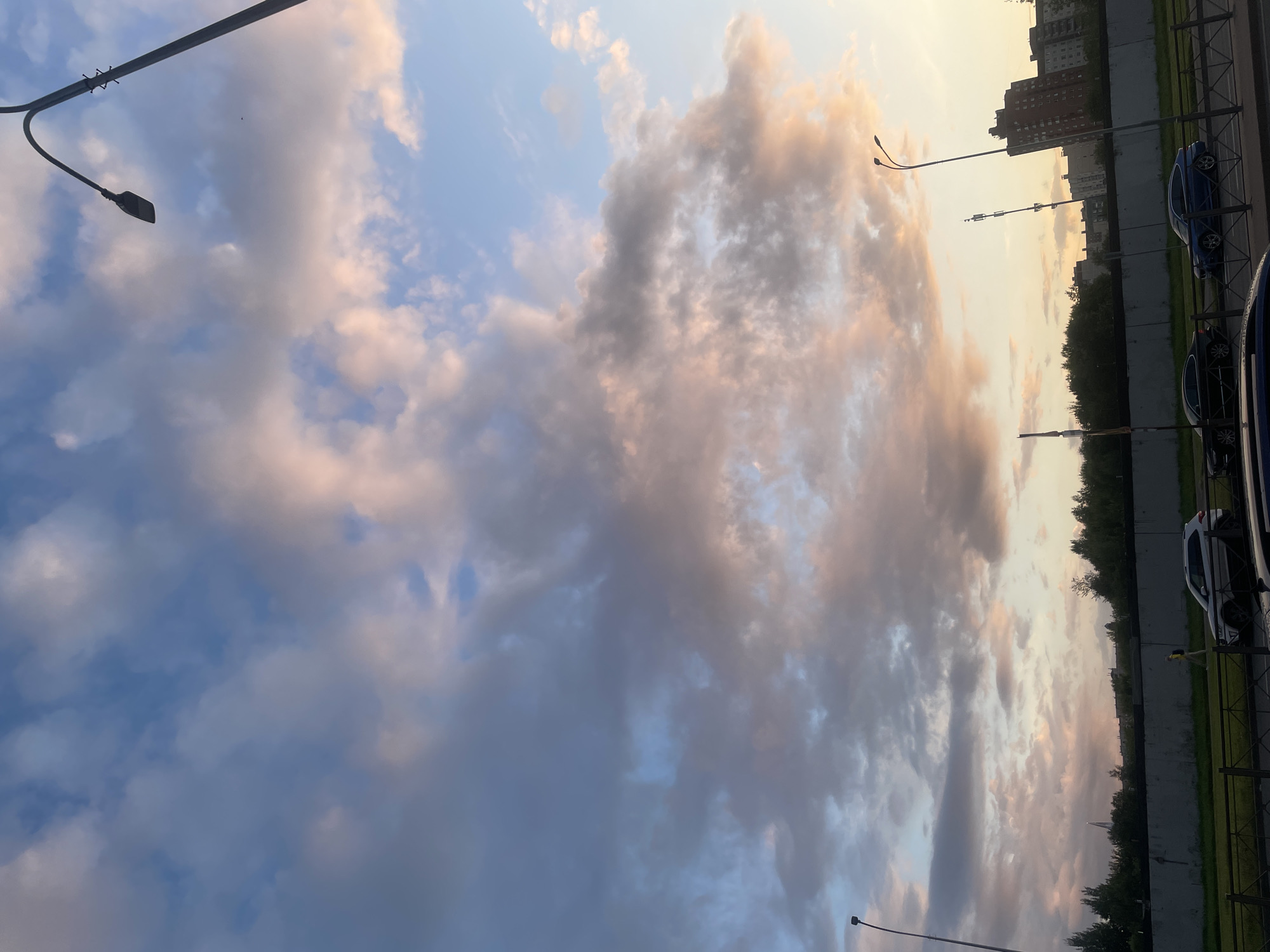 The image shows a street view in Saint Petersburg, Russia, during a cloudy sunset. The sky is filled with dramatic clouds tinged with pink and orange hues, contrasting with the deep blue sky. A street lamp with a curved arm stands prominently in the foreground on the right. In the middle ground, a row of trees lines the horizon, with several buildings visible behind them. A concrete barrier runs along the street, and a metal fence separates the sidewalk from the road. Several cars are parked along the street, including a white SUV, a black sedan, and a blue sedan. A person is walking on the sidewalk. The scene captures an urban environment under a colorful, atmospheric sky.