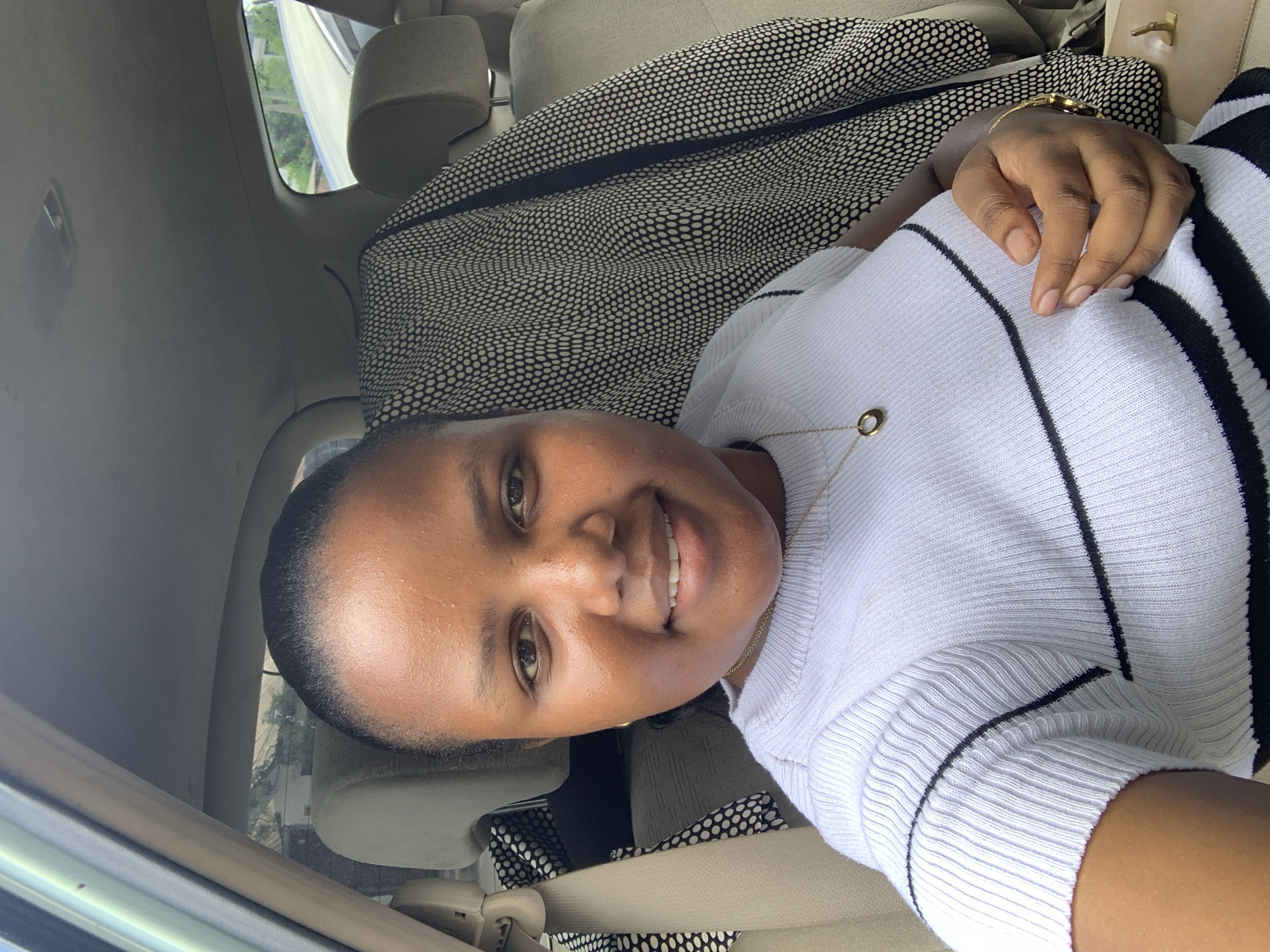 A person with dark skin and short, dark hair is in a car, appearing to be in Dar es-Salaam, Tanzania. The person is wearing a white shirt with black stripes. They are smiling and looking at the camera. Their hand is visible, wearing a gold bracelet. A black and white patterned bag is behind them. The interior of the car is light-colored. The location, Dar es-Salaam, is inferred from the visible buildings and vegetation outside the car window.