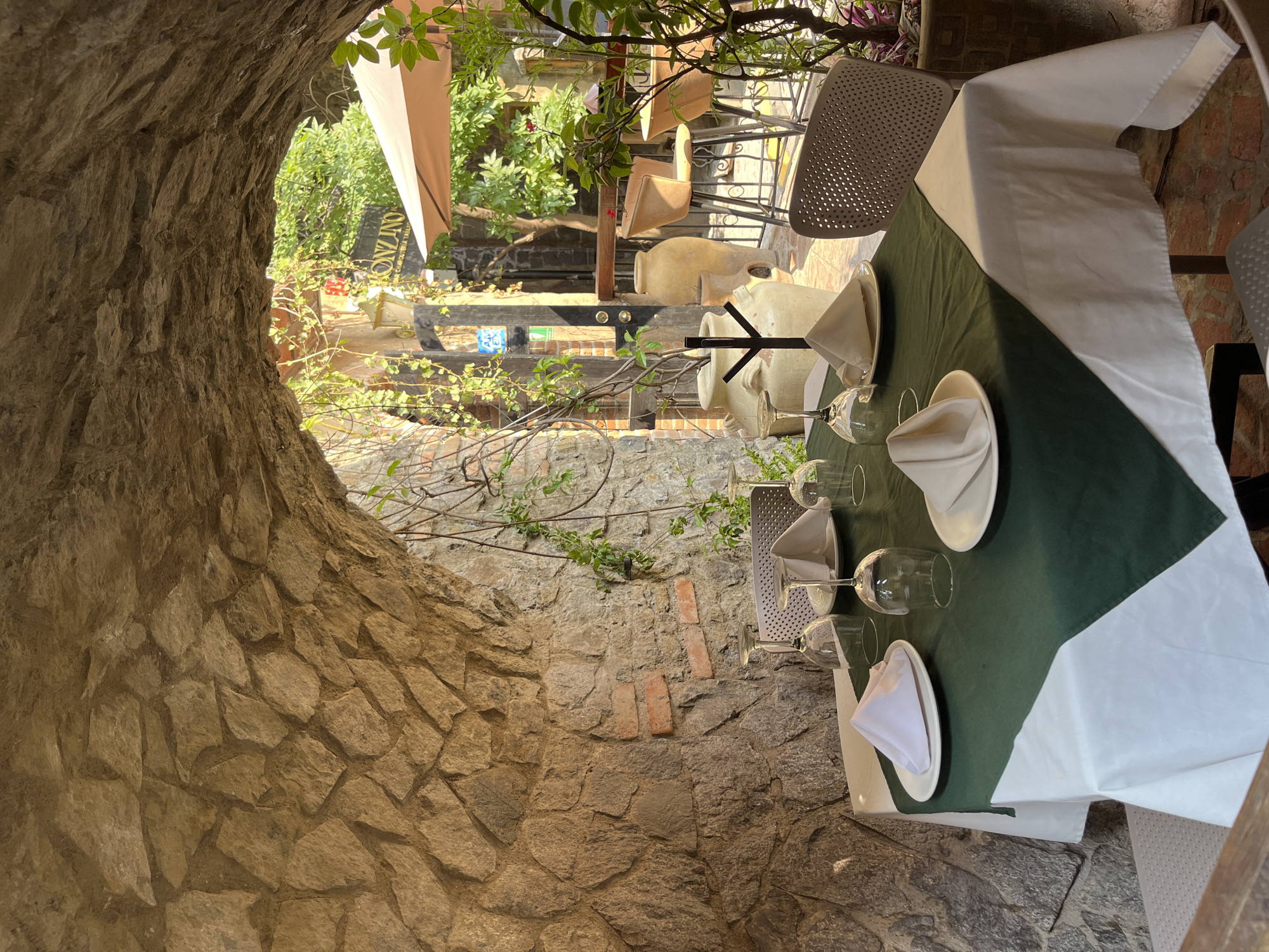 The image shows a set table at an outdoor restaurant in Domingo Arenas, Mexico. The table is covered with a green and white tablecloth and set with plates, napkins, and wine glasses. The restaurant is located in a stone structure with an arched opening that looks out onto a courtyard. The courtyard contains additional tables, chairs, and plants. A sign for the restaurant is visible in the background. There are no people visible in the image.