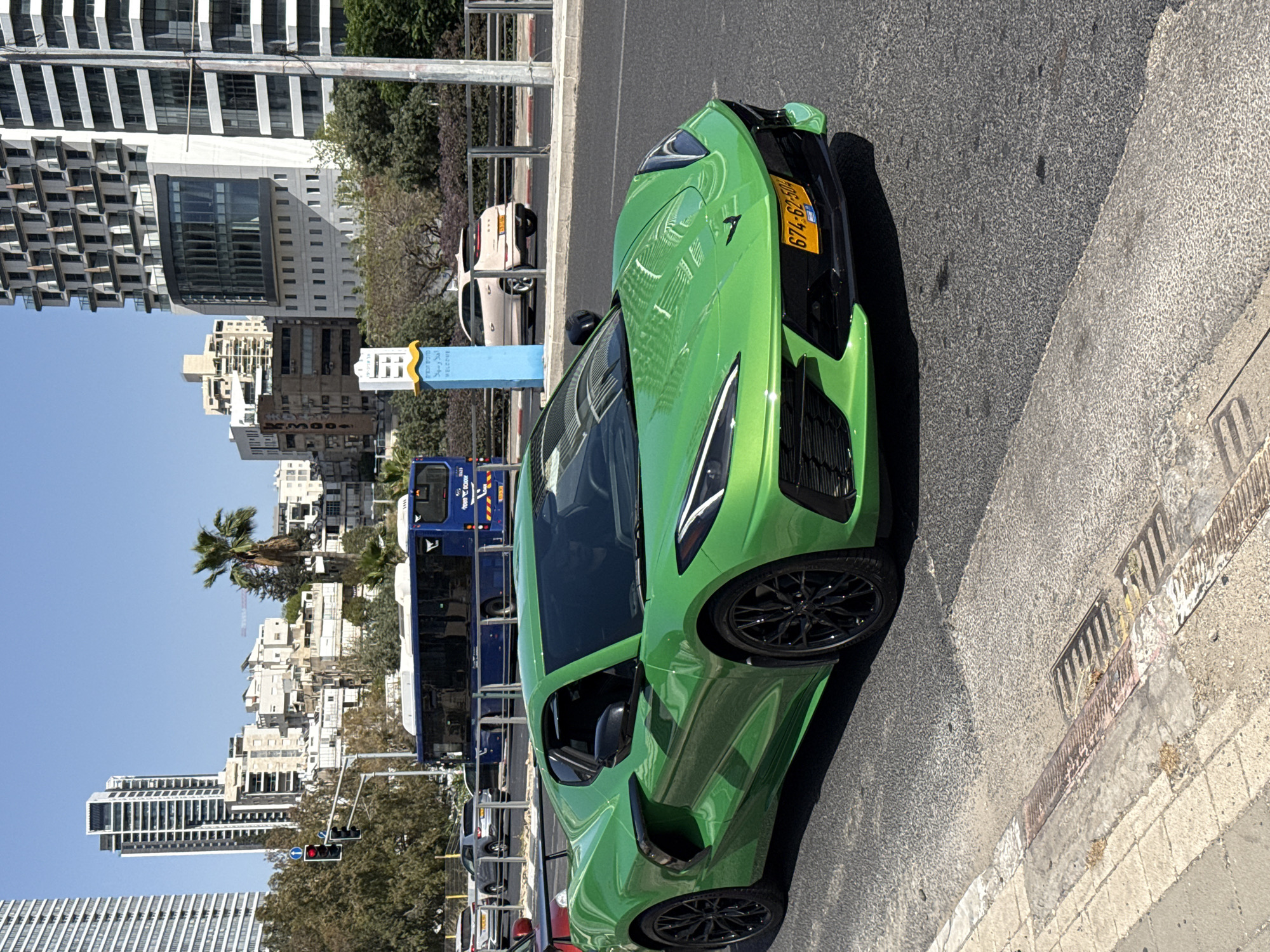 A green sports car is parked on the right side of a street in Tel Aviv, Israel. The car's license plate is visible, displaying the number 674-62-749. Behind the car is a blue bus, and in the background, there are buildings that make up the city skyline. There is a palm tree to the left of the frame, and the sky is clear and blue.