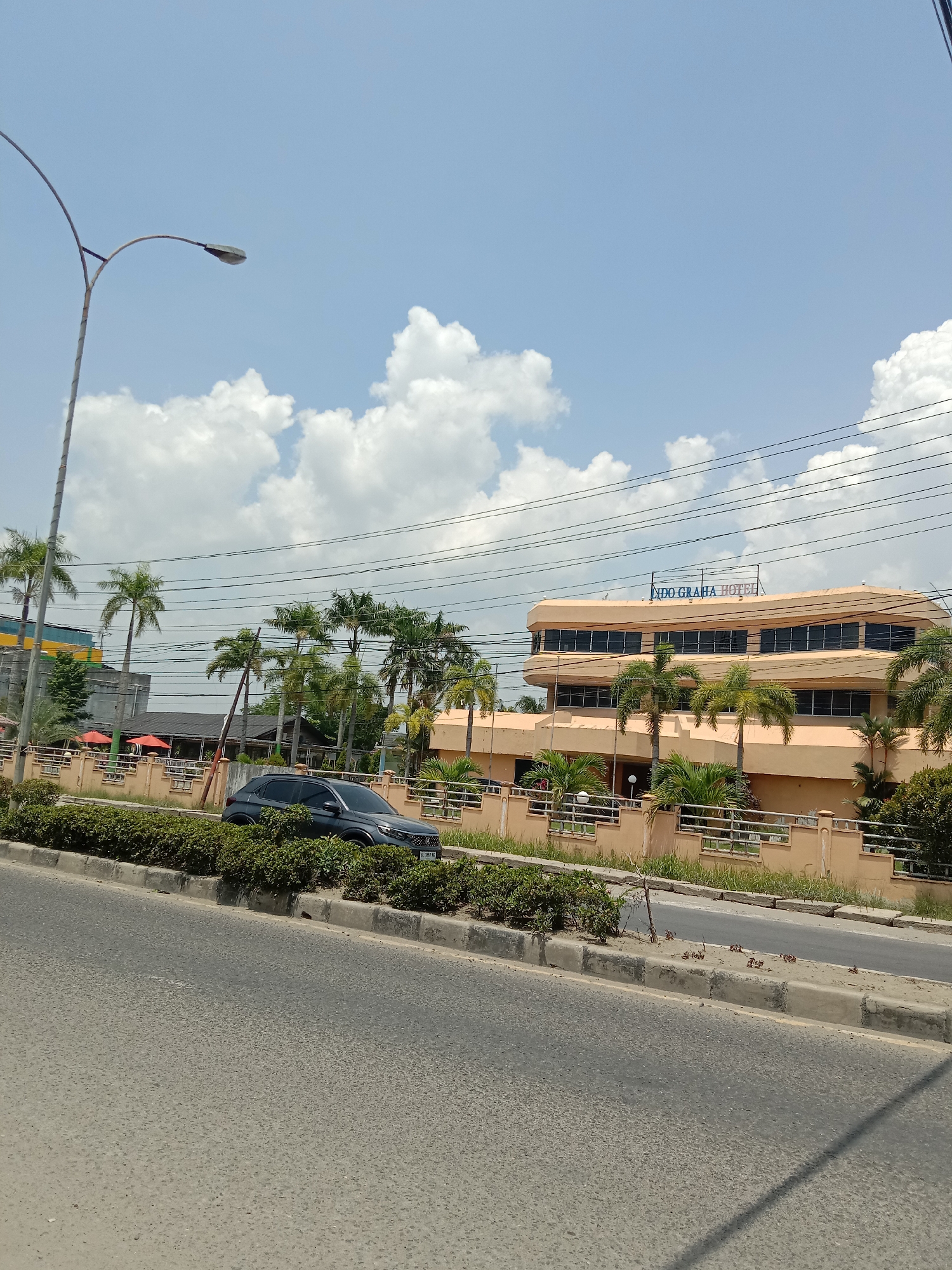 The image captures a daytime street scene in Lhokseumawe, Indonesia, under a partly cloudy sky. The main subject is a large, light orange-colored building identified as "INDO GRAHA HOTEL," with its name prominently displayed in blue letters on the top facade. The hotel features multiple floors with horizontal window bands and a somewhat curved, modern architectural design.
In the foreground, a paved road stretches across the bottom of the frame, separated from the hotel premises by a concrete curb and a landscaped median filled with green bushes. A dark-colored car, possibly an SUV or hatchback, is parked or slowly moving behind these bushes, facing slightly to the right.
Numerous tall palm trees are scattered around the hotel property, particularly in front and to the left of the building, enhancing the tropical setting. Power lines crisscross the upper middle section of the image. On the far left, a tall street lamp with a curved arm is visible, and in the distance, other buildings with varying structures can be seen, partially obscured by trees.
The sky is bright blue with large, fluffy white cumulus clouds, indicating a clear and sunny day. No people are visible in the frame, and there isn't a specific event taking place beyond the general activity of a street scene. The image provides a snapshot of an urban environment with a prominent hotel and natural greenery.