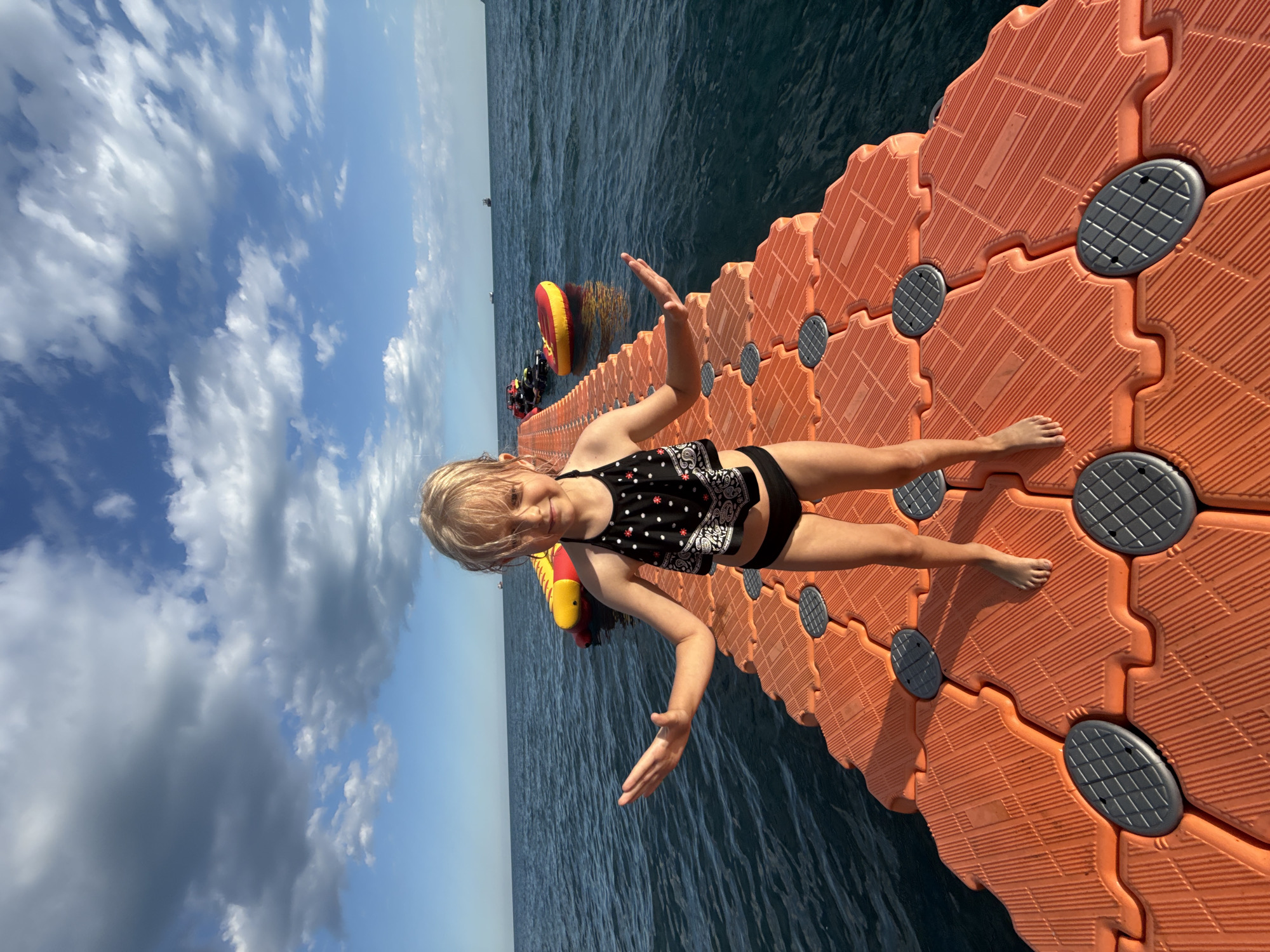 A young girl with blonde hair and a black and white polka dot bikini stands on an orange floating dock. She is facing the camera with her arms outstretched, palms up. The dock is made of interlocking hexagonal modules and extends into dark blue water. In the background, a red and yellow inflatable object floats on the water. The sky above is blue with fluffy white clouds. The sun is shining, indicating it is daytime. The scene appears to be a recreational area on the water, possibly a lake or the sea, near Yalta, Ukraine. The girl looks happy and excited. There is no visible text in the image.