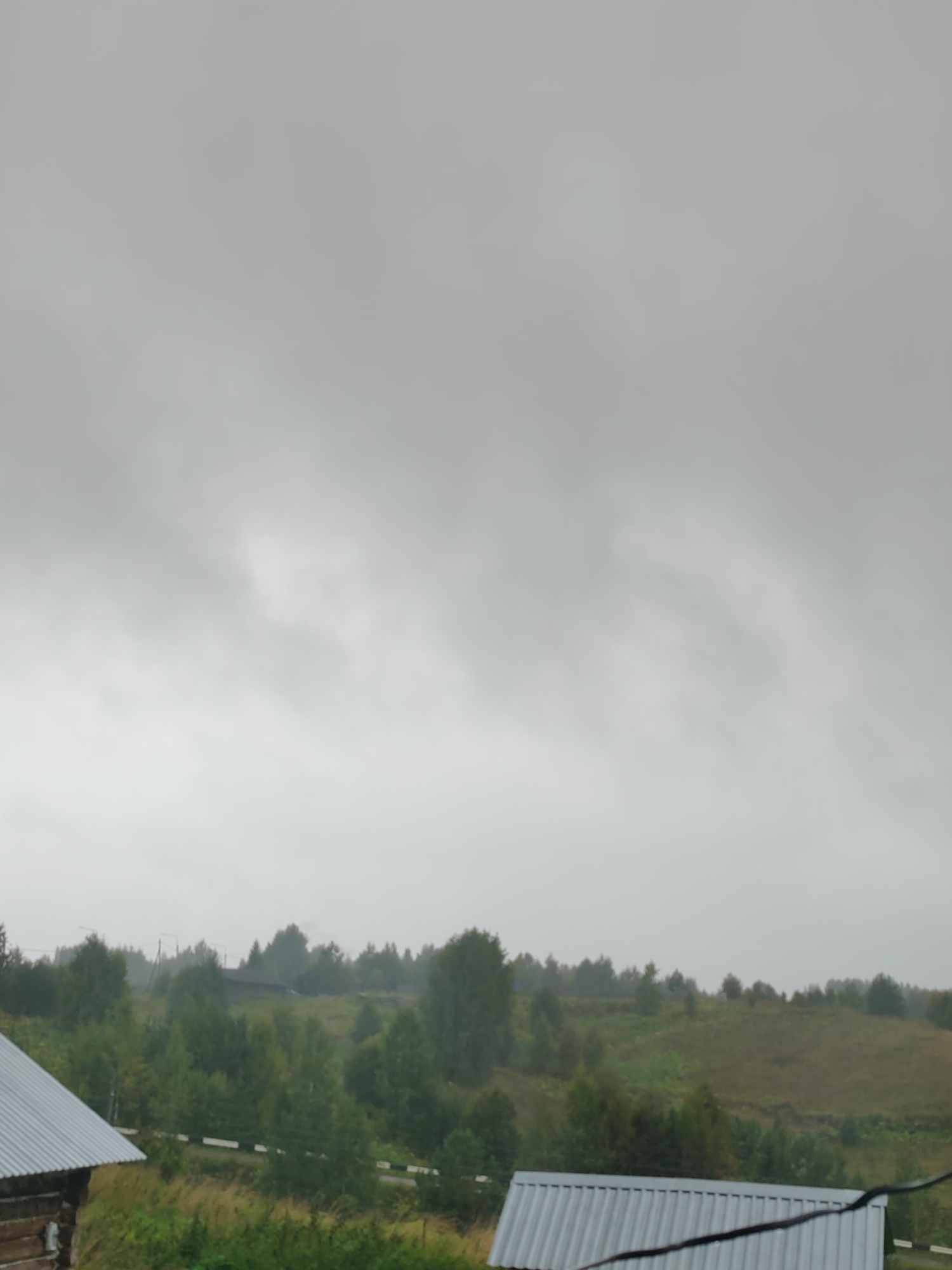 The image depicts a landscape under an overcast, gray sky, suggesting a cloudy or foggy day. In the foreground, the corrugated metal roof of a building is visible on the left, and a similar roof structure extends across the bottom right of the frame. The midground is characterized by a rolling, green hill dotted with trees of various sizes and densities, some appearing as deciduous foliage and others as evergreens. A road with white and black striped markings runs horizontally across the midground, separating the grassy area from the tree line. In the distance, the landscape continues with more trees and hints of human habitation, possibly a small village or scattered houses, indicated by darker shapes amongst the foliage and faint electrical poles. The overall impression is a rural scene, likely in a temperate climate, with a focus on natural elements and minimal signs of human activity. The location is specified as Chukhlem, Russia.