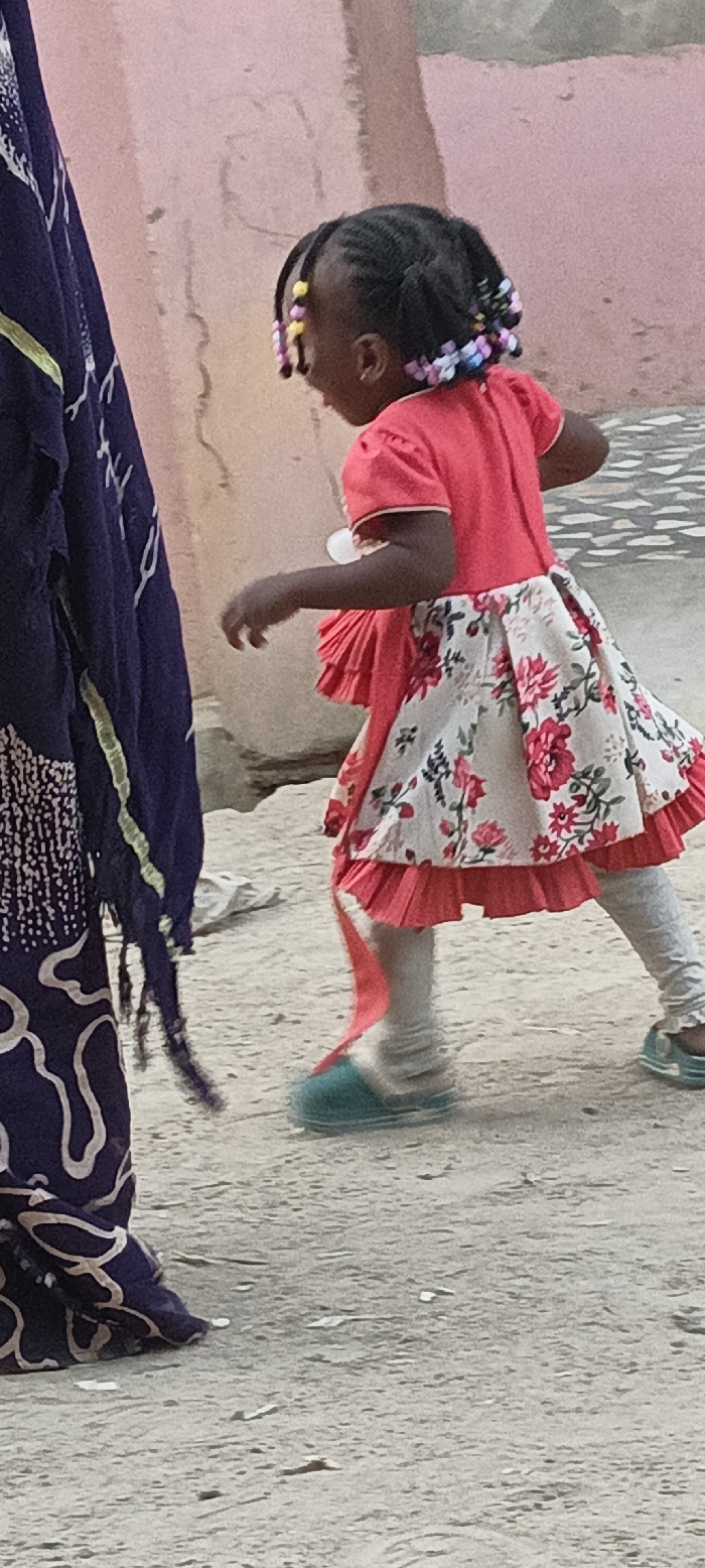 This vertical full shot depicts an outdoor scene in Rangaza, Nigeria, featuring a young child in motion, viewed from behind and slightly to the right, with a partially visible adult on the left. The child, likely a girl, wears a bright red, short-sleeved top and a patterned skirt that is predominantly white or cream with bold red and green floral designs. Underneath the skirt, red pleated ruffles are visible. She has light grey leggings and turquoise or teal slippers on her feet, with the left foot appearing blurred, suggesting movement. Her dark hair is styled in numerous braids, adorned with colorful beads in yellow, pink, purple, and light blue.
To the left of the frame, the lower body and midsection of an adult are visible, clad in a dark blue fabric with intricate white or light-colored swirling patterns and fringes, possibly a traditional garment. The background consists of a plain, light pinkish-brown wall, suggesting a building, while the ground is sandy or dusty. The natural lighting indicates it is daytime, likely morning or late afternoon. The overall setting and attire are consistent with a local environment in Rangaza, Nigeria. No specific emotions are discernible as faces are not clearly visible, and there is no visible text in the image.
