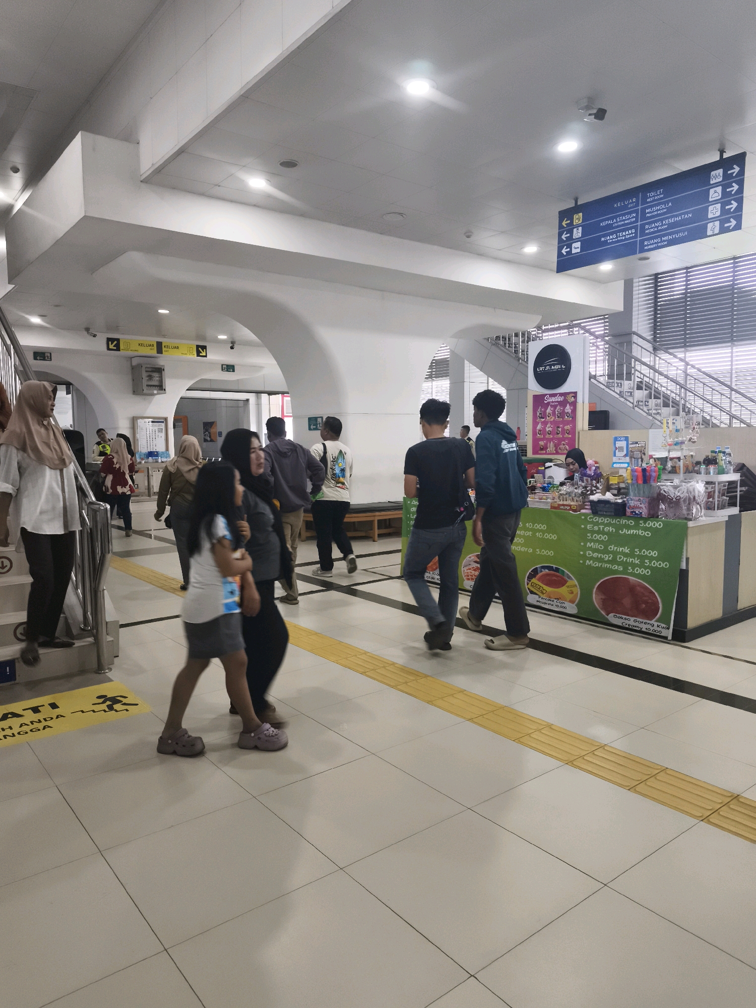 The image depicts an indoor public space, likely a train station or a similar transportation hub, given the presence of directional signage. The environment is well-lit with fluorescent ceiling lights and appears to be clean and modern. The floor is tiled in a light color with a yellow tactile paving strip running horizontally across the scene, indicating a pathway.
In the foreground, a young girl wearing a white top and a gray skirt, along with pink clogs, is walking towards the left, her body angled away from the viewer. To her right and slightly behind her, an adult woman wearing a dark hijab and a dark top is walking in the same direction, looking towards the right.
Further back, on the left side of the image, a woman wearing a beige hijab and a white long-sleeved shirt is descending a set of stairs. Another woman in a red and white patterned top is walking away from the stairs towards the center of the image.
In the middle ground, a group of people are walking through an arched entryway. Two men, one in a black t-shirt and jeans and another in a dark blue hoodie, are walking towards the right. Behind them, a man in a grey hoodie and dark pants is walking towards the left. Another man in a white t-shirt with a graphic design is walking towards the right.
To the right of the image, a food stall with a green banner displaying various food and drink items and their prices is visible. A person with a dark hijab is attending the stall. The banner lists items like "Cappuccino 5.000", "Es Teh Jumbo 5.000", and "Bakso Goreng Kuah Creamy 10.000". Behind the stall, a staircase with metal railings ascends to an upper level.
Overhead, a blue directional sign hangs from the ceiling, providing directions to various facilities including "KELUAR" (Exit), "TOILET", "KEPALA STASIUN" (Station Master), "MUSHOLA" (Prayer Room), "RUANG TENANG" (Quiet Room), and "RUANG KESEHATAN" (Health Room). Another set of yellow directional signs with arrows pointing left and right are visible on the wall above an arched opening.
The overall scene suggests a busy transit environment with people moving through the space and patronizing the food stall. The location is identified as Kelurahan 16 Ilir, Kecamatan Ilir Timur I, Indonesia.