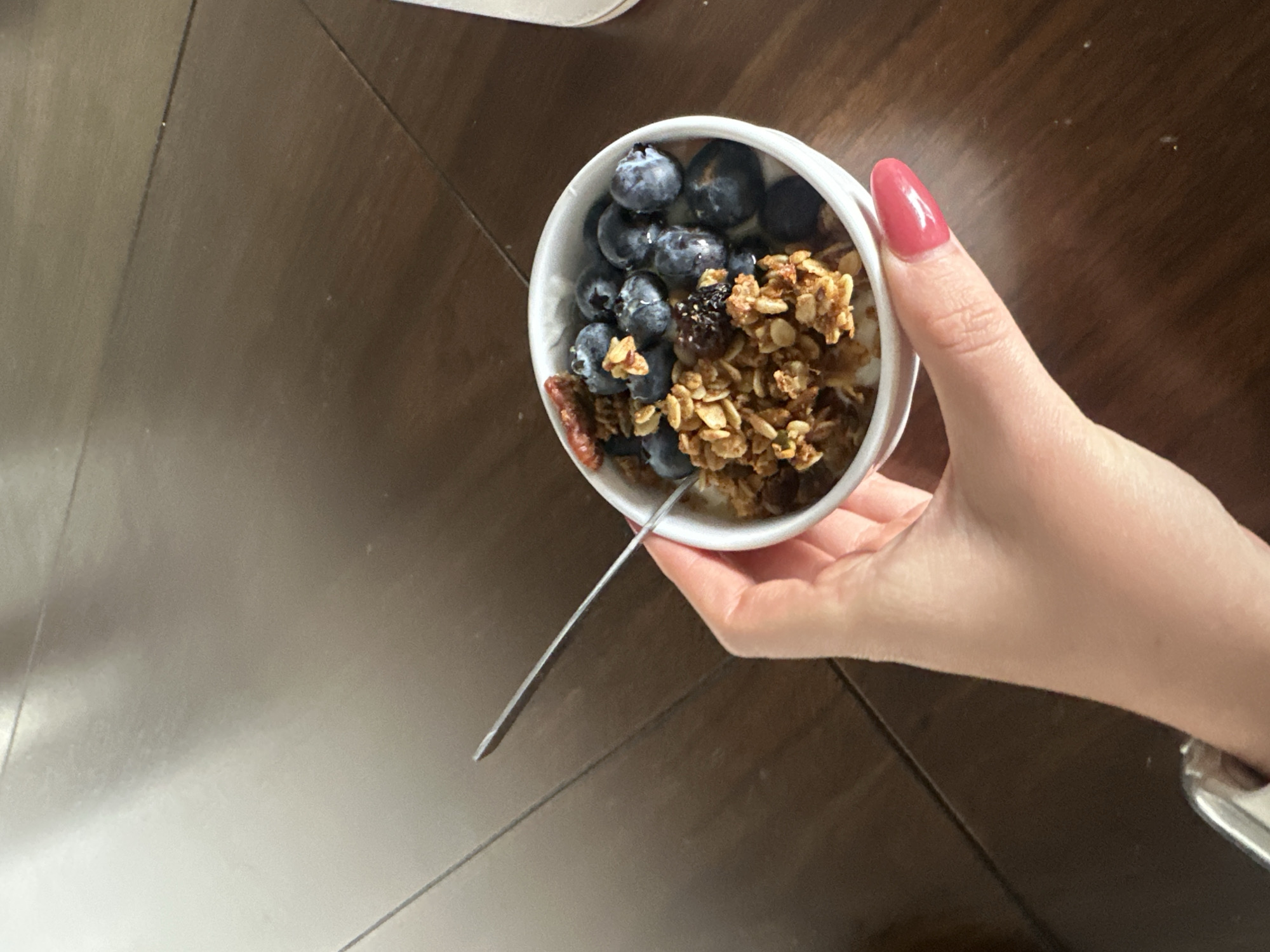A hand holds a white bowl filled with yogurt, blueberries, and granola over a wooden surface. A spoon is visible in the bowl.
There is no information to indicate that the location is in Huixquilucan de Degollado, Mexico.