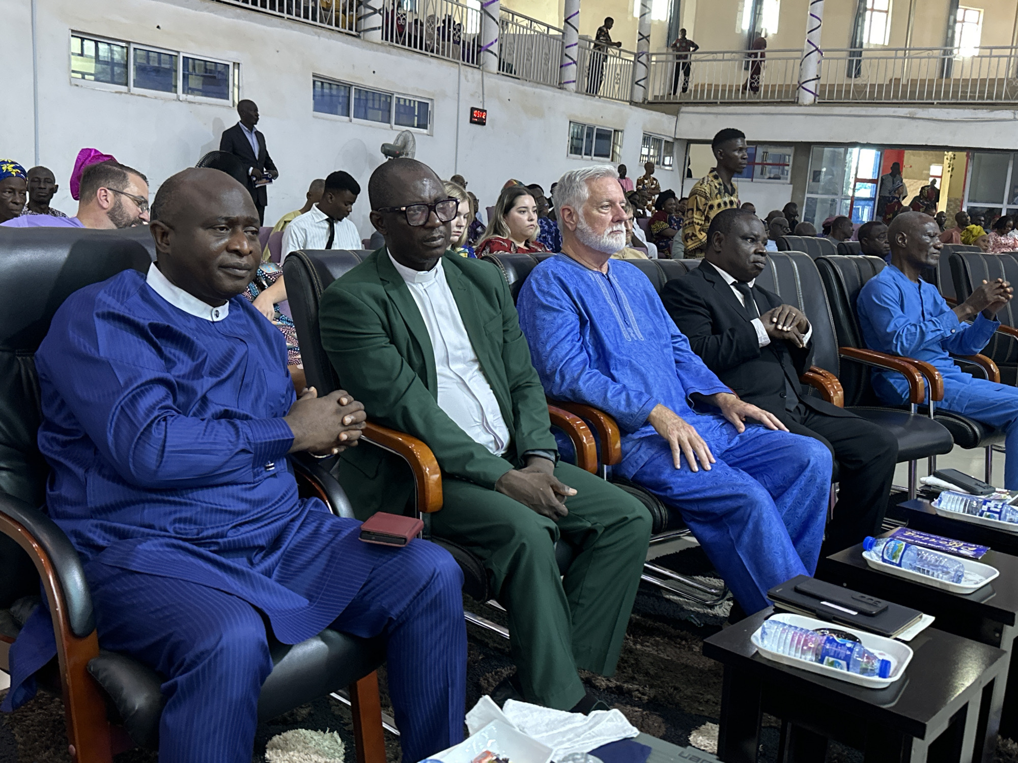 In a large indoor venue, likely a church or conference hall, a group of men and women are seated in chairs arranged in rows. The scene is lit by artificial lighting. The overall atmosphere appears to be one of a formal gathering or service.
In the foreground, three men are prominently featured, seated next to each other. The man on the left, wearing a blue traditional attire, is facing forward with his hands clasped in his lap. The man in the middle, in a green suit and white shirt, is also facing forward, looking slightly to his left. He has glasses on and is wearing a clerical collar. To the right of the man in green is a man with a white beard, wearing a blue traditional outfit. He is also facing forward.
Behind these men, other attendees are visible, some looking towards the front, others engaged in conversation. Some individuals are wearing traditional African clothing, while others are in Western attire. The background shows architectural features of the venue, including columns, balconies, and windows. A digital clock displaying "2:51" is visible on the wall in the upper middle section of the image.
On the tables in front of the seated individuals, there are trays with bottled water and what appear to be wrapped snacks or candies. Mobile phones are also visible on some of the tables. The flooring appears to be carpeted. The setting suggests a gathering in Television, Nigeria, possibly a religious or community event.