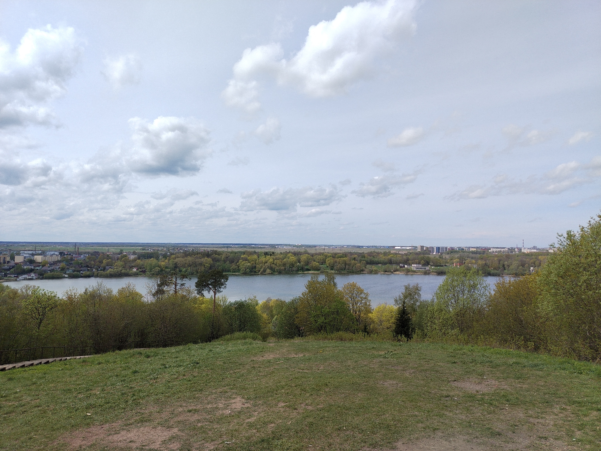 The image shows a landscape view overlooking a body of water, likely a lake or wide river, from a grassy hill. The sky is overcast with grey and white clouds.
In the midground, a line of trees separates the water from the background. On the opposite shore of the water, buildings and structures characteristic of an urban area are visible.
The foreground is composed of a sloping grassy hill with patches of bare earth. Some shrubs and trees are scattered across the hill.
The image captures a view from a natural elevation point overlooking the urban landscape and a water feature in Krasnoye Selo, Russia.