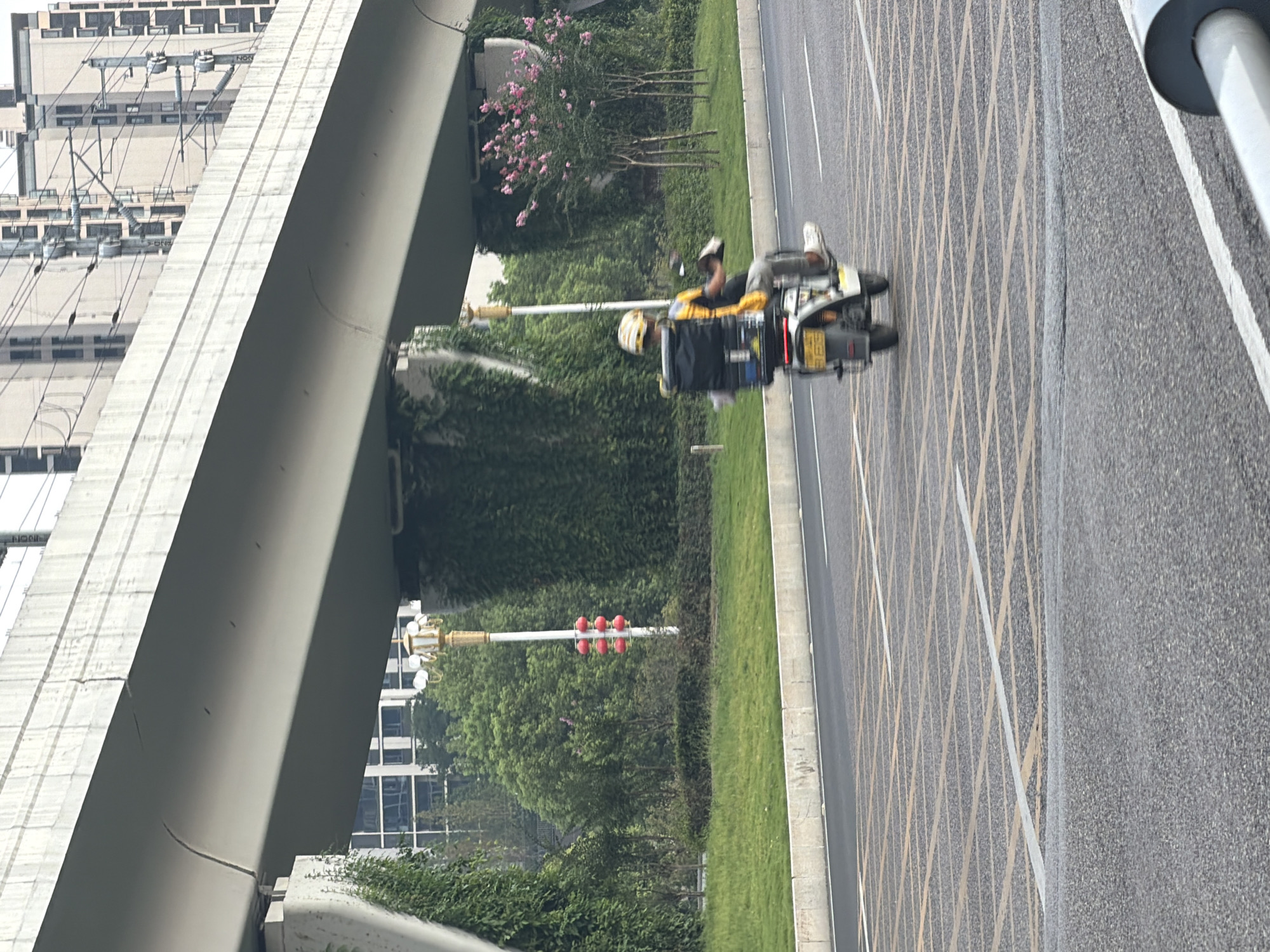 A person is riding an electric scooter with a large black delivery box on the back, on a road in Jiangjin, China. The scooter is white with some yellow accents and a yellow license plate with Chinese characters and numbers. The rider is wearing a helmet and a yellow and black jacket. They are on a road with yellow grid markings and white lane lines. In the background, there is a large elevated concrete structure, possibly an overpass or elevated train line, with buildings visible behind it. Green trees and shrubs line the side of the road, and there is a lamppost with red spherical decorations. The sky is overcast.