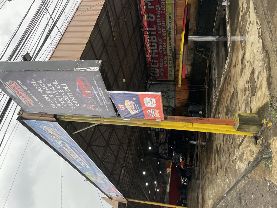 The image shows a car wash facility in Ngamprah, Indonesia, located at latitude: -6.863, longitude: 107.504. The facility is situated under a metal awning and has a concrete floor that is wet and appears to be recently cleaned. Several cars are parked inside, and one is being serviced by a worker. The facility is advertised as a "Cuci Mobil & Motor", offering services such as interior and body cleaning, car detailing, and engine maintenance. The company's name is "Sultan Car Wash", and its contact information is displayed on a sign. There are also several other signs, some of which are in Indonesian, promoting the facility's services. The surrounding environment is typical of a suburban neighborhood in Indonesia, with concrete buildings and paved roads. The image was taken on a cloudy day, with no visible sunshine. The overall impression is of a typical, busy car wash facility in a developing country.