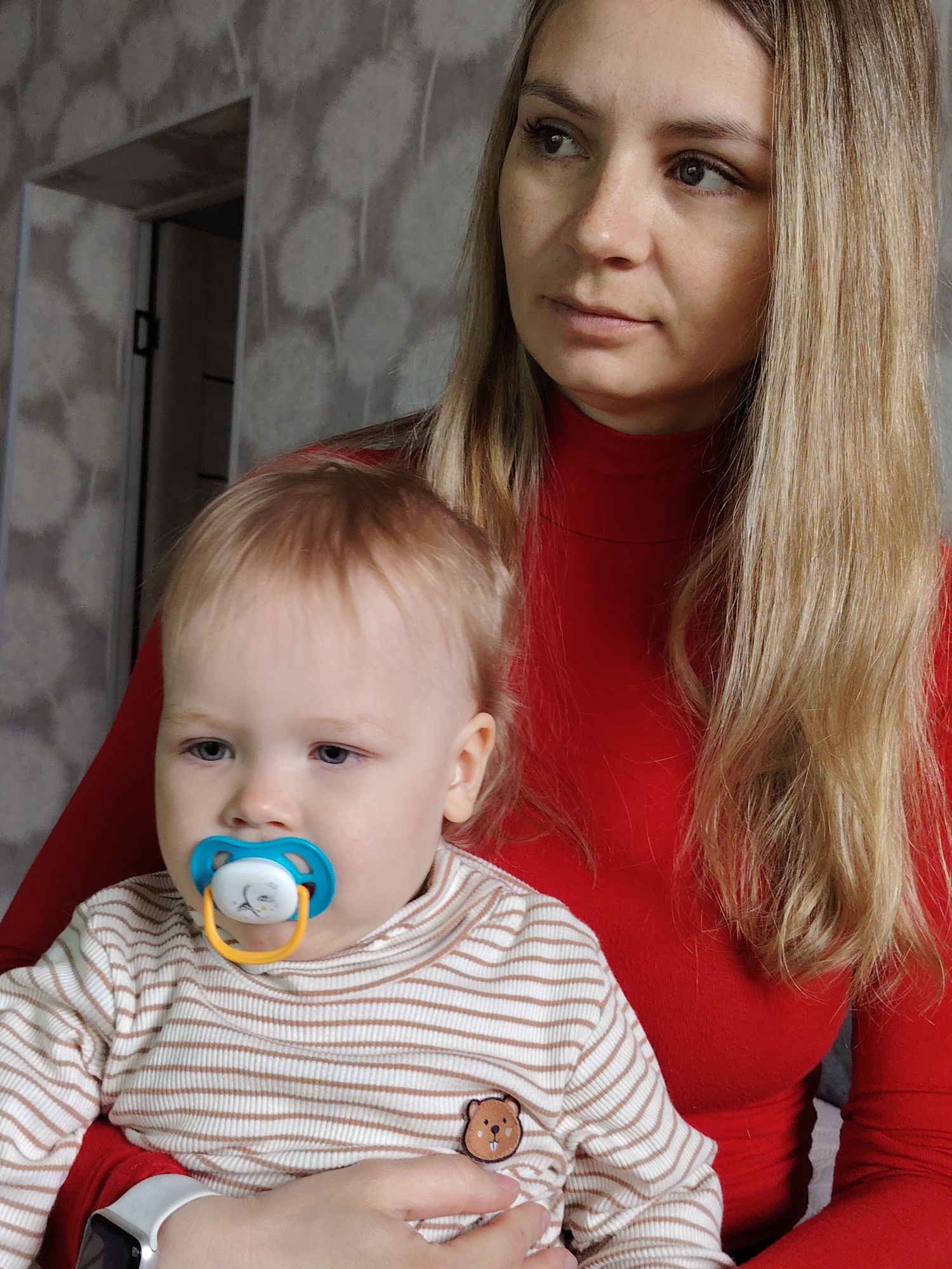 A woman with long blonde hair, wearing a red turtleneck sweater, is holding a baby in her arms. The baby, who has fair hair and blue eyes, is wearing a striped top with a small bear patch. The baby has a blue and white pacifier in its mouth. The setting appears to be indoors, with a patterned wall visible in the background and a doorframe slightly to the left. A watch is visible on the mother's wrist. The image was taken in the city of Chaikovsky, Russia.