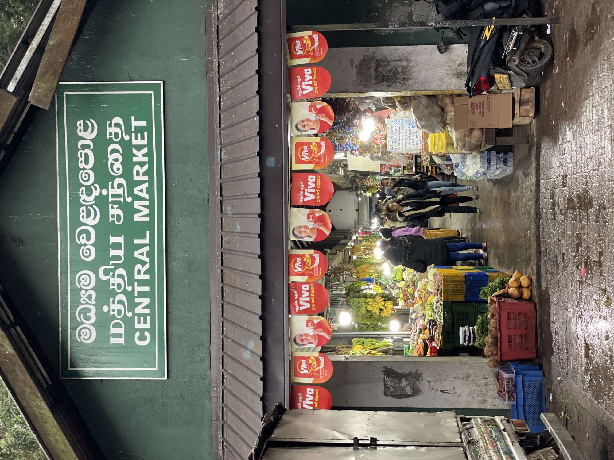 The image shows the entrance to the Central Market in Nuwara Eliya, Sri Lanka. A green sign displays the market's name in multiple languages. Above the entrance, red and white "Viva" advertising banners are affixed. Inside, a bustling market scene is visible with people shopping among displays of fruits and vegetables stacked in crates. A motorbike is parked outside the market. The ground appears wet, possibly from recent rain.