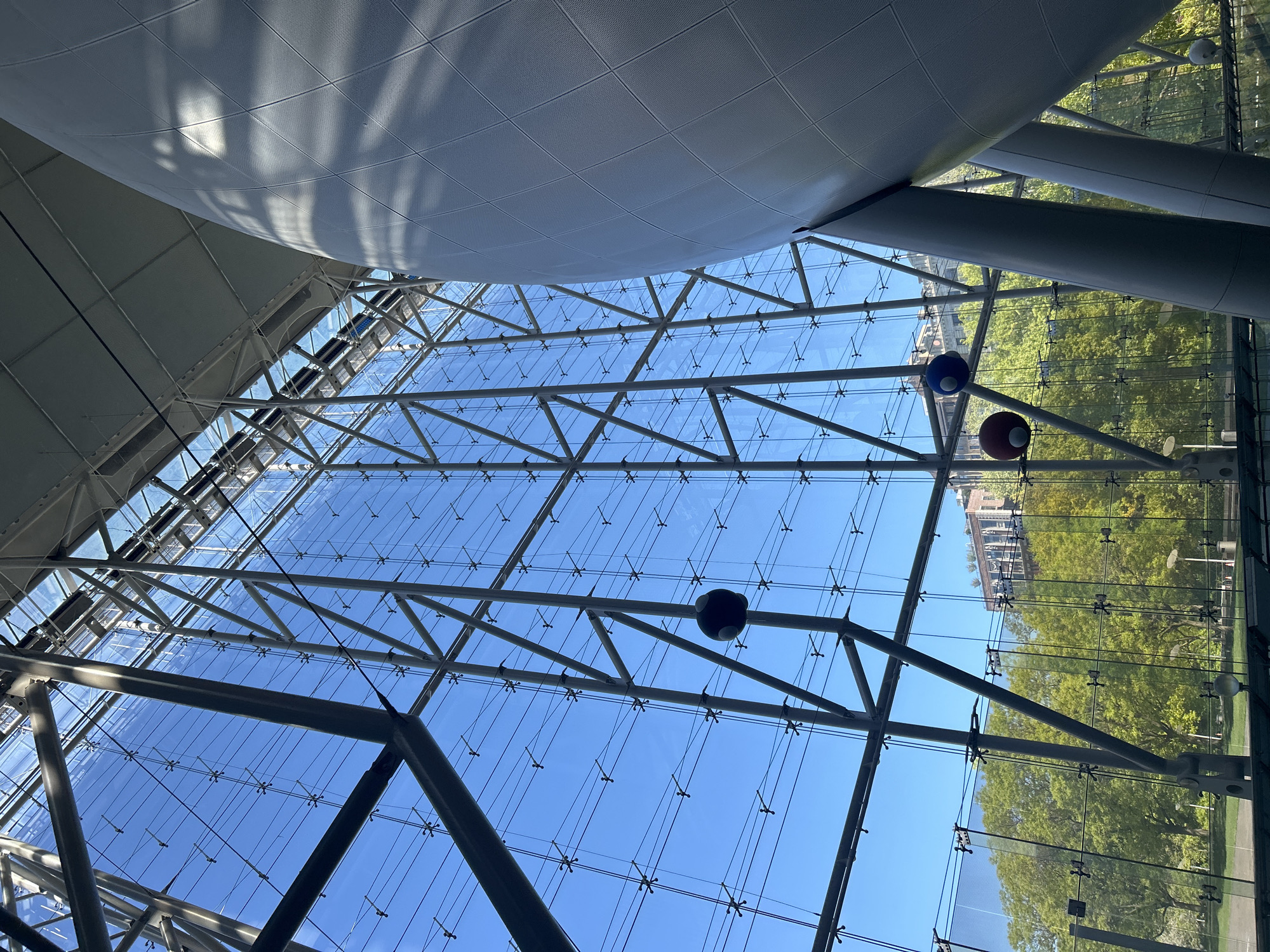 The image captures an upward-looking perspective inside a contemporary building, dominated by an expansive glass and metal ceiling structure. A clear, bright blue sky is visible through the transparent glass panels, which are held in place by numerous small, star-shaped connectors and supported by a robust network of grey metal beams and trusses forming intricate geometric patterns.
In the upper section of the frame, a large, curved, white ceiling element, possibly made of perforated metal or fabric, casts distinct shadows from bright sunlight, indicating a clear daytime setting. Several spherical, decorative objects are suspended from the ceiling structure; prominent examples include one blue and one red sphere visible on the right side, and another dark grey/black sphere further left.
Beyond the glass roof, the environment appears to be a vibrant outdoor space. Lush green trees, likely part of a park or large garden, are visible, bathed in sunlight. In the distance, fragments of urban architecture, including multi-story buildings with detailed facades, can be seen, suggesting an urban context. The overall impression is one of architectural grandeur, openness, and abundant natural light, characteristic of modern public or commercial spaces in a city like New York, United States of America. No specific human activity or visible text is present.