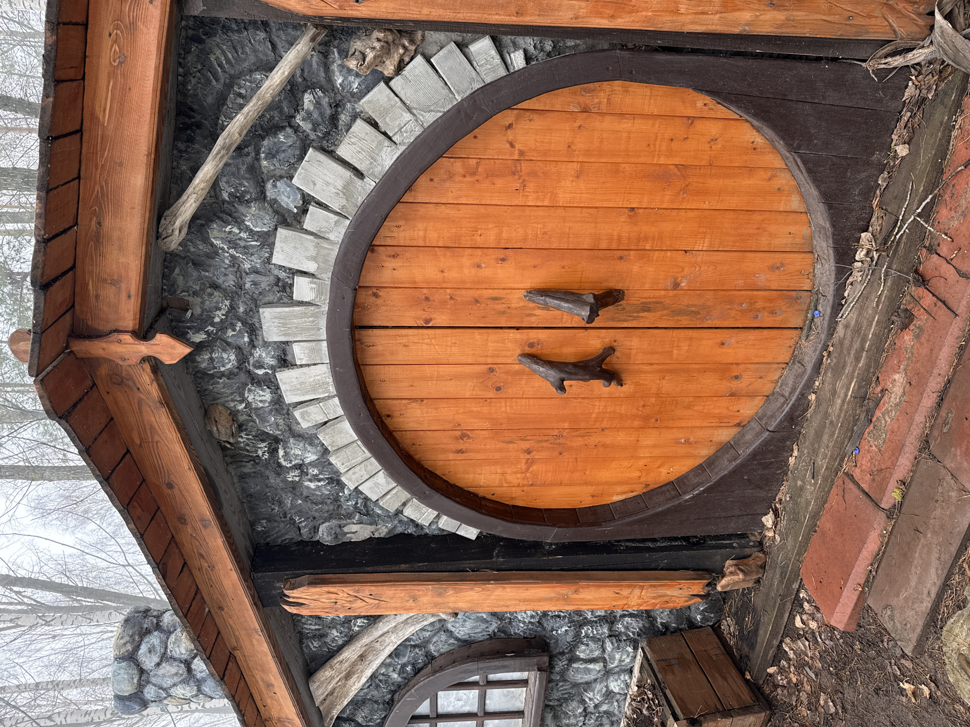 The image shows a close-up of a whimsical, round wooden door set into a building facade constructed with gray stones and gray brick accents. The door has two stick-like handles. The building’s wooden framing, including roof overhangs, is visible. The environment appears to be outdoors, possibly a yard or garden. Based on the location, this could be a decorative element within a recreational or park area of Chaikovsky, Russia.