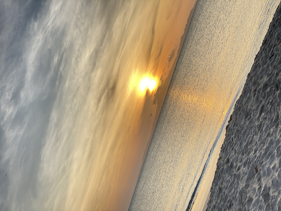 A rotated seascape image shows a beach where sunlight reflects off the water, creating a bright streak. The beach sand is textured. The sky transitions from gray-white clouds to a golden hue near the horizon, where the sun shines brightly. The clouds are striated, adding a sense of movement to the serene scene.
