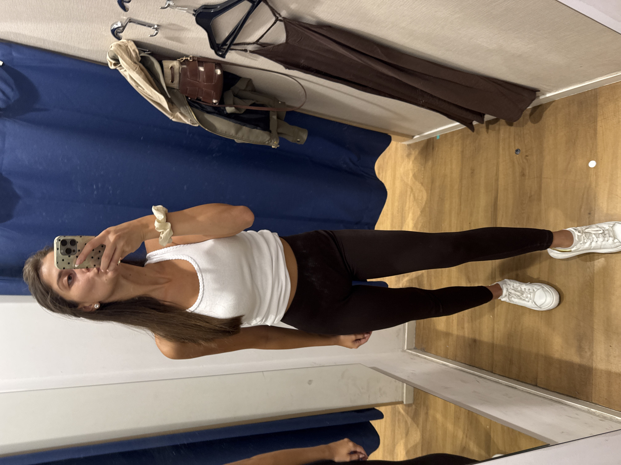 A young woman is standing in what appears to be a clothing store fitting room, taking a mirror selfie with her smartphone. She is wearing a white tank top, dark leggings, and white sneakers. The fitting room has a blue curtain on one side and wooden flooring. Clothes are hanging on a rack. There is a mirror showing the reflection of the lower part of her body. The location in the city of Burgas, Bulgaria is not visible or verifiable in this image.
