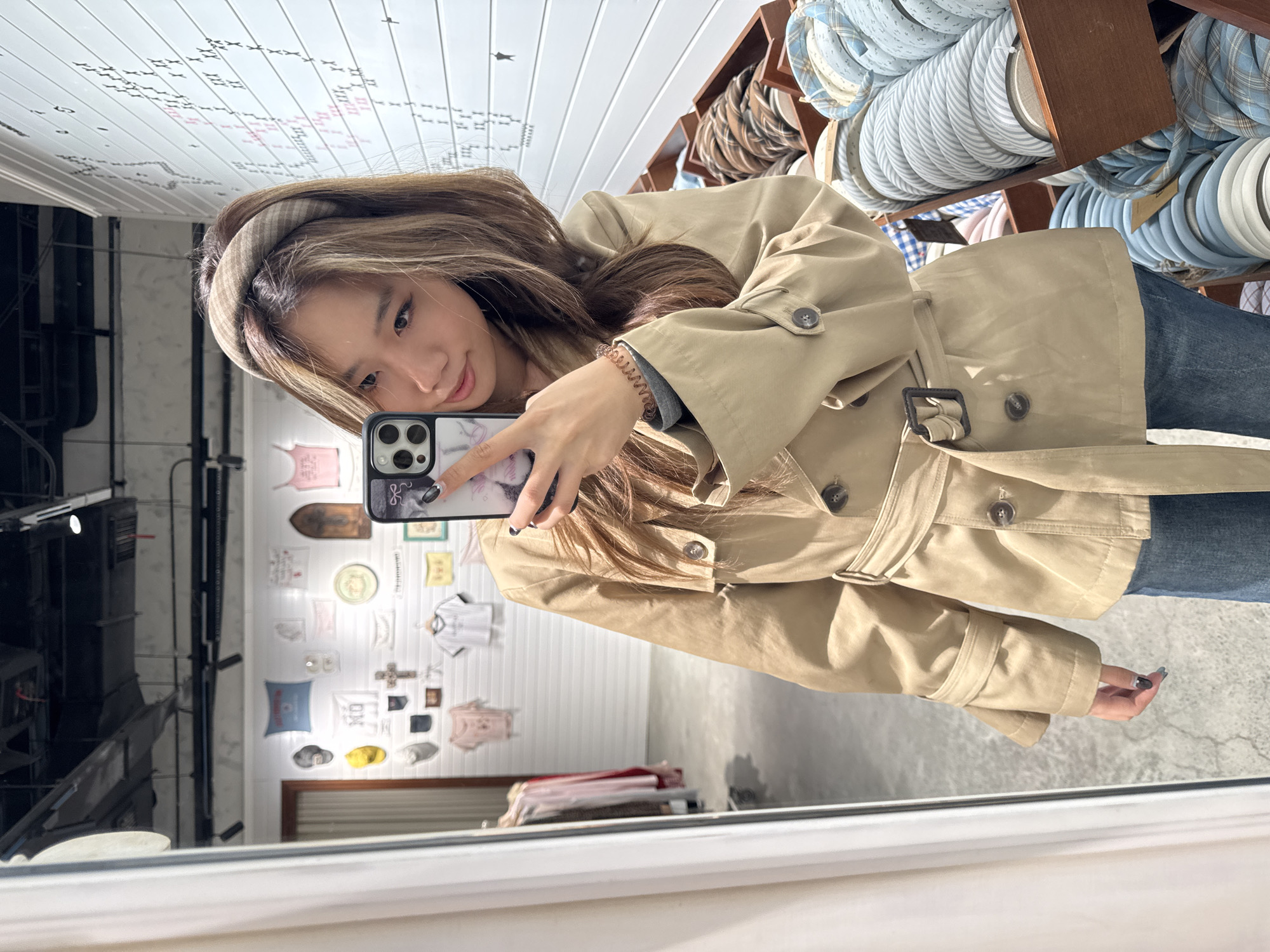 A woman takes a selfie in a mirror. She wears a beige trench coat and a patterned headband. A smartphone is in her hand. The background behind her in the mirror shows a wall adorned with various decorative items and clothing. Above her is a display of stacked rolls of fabric. The reflection in the mirror shows a modern interior. Jianlan Road District, China is not apparent from the image.