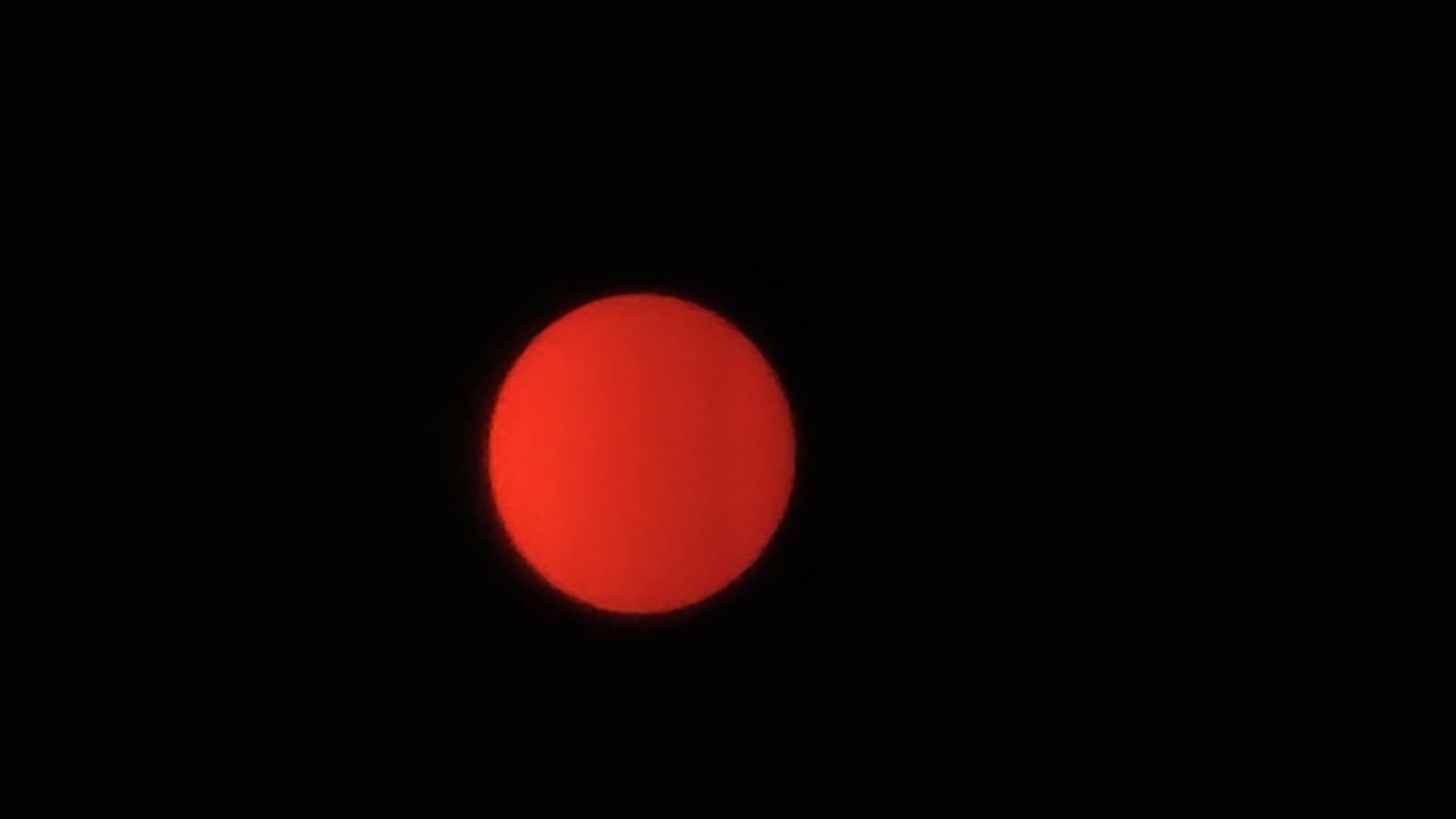 The image features a prominent, intensely red, circular object set against a completely black background. The red circle is large and centrally positioned, appearing as a solitary light source. Its vibrant color strongly suggests a celestial body, such as the sun or moon, observed during sunrise or sunset, with its light being heavily filtered by atmospheric conditions like haze, smoke, or dust. The edges of the red circle appear slightly soft, indicating possible atmospheric diffusion or a zoomed-in perspective. The background is uniformly dark, entirely devoid of any other visual details such as stars, clouds, or terrestrial features. There are no visible people, objects, or text within the frame, creating a stark and minimalist composition. The scene captures a powerful natural phenomenon in what is identified as the Bac Natural Heritage Area in Trang, Vietnam.