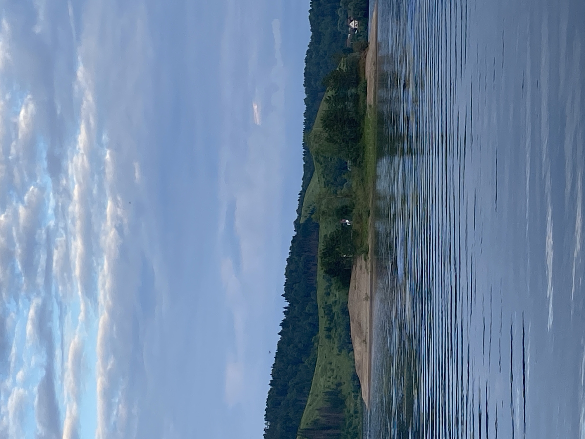 This is a vertical, full shot of a tranquil natural scene in Noviy, Russia, likely captured during the late afternoon or early evening. The image is divided into two primary sections: the sky and the landscape bordering a body of water.
The left side of the frame is dominated by a vast, pale blue sky filled with scattered, wispy white clouds. The clouds are softly illuminated, suggesting a gentle light, possibly from a setting sun or overcast conditions.
The right side of the image presents a serene landscape with a body of water in the foreground. The water's surface is disturbed by numerous parallel ripples, creating a textured, reflective effect that mirrors the sky. The water appears calm and deep blue. Adjacent to the water is a sandy bank, which then gives way to a lush, green hillside covered in dense forestation. The trees are a dark, verdant green, creating a strong contrast with the lighter sky and water. A few scattered buildings, appearing to be houses with white roofs, are visible nestled on the top of the hill, hinting at a rural or semi-rural settlement.
There are no people or specific objects of interest clearly depicted in the scene, emphasizing the natural beauty and peaceful atmosphere. The overall mood is one of quietude and natural serenity. No text is visible in the image.