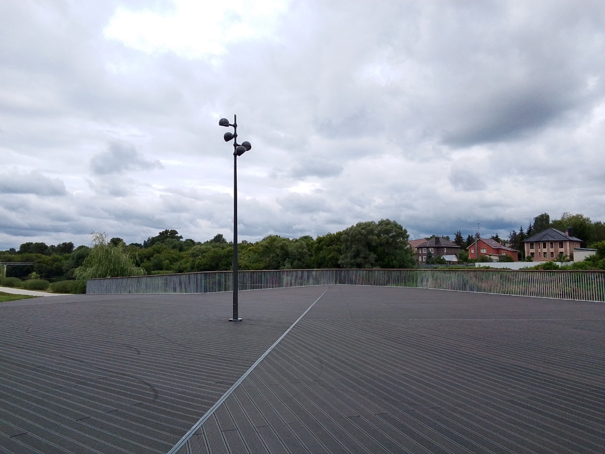 The image depicts an outdoor scene on a cloudy day, likely in the afternoon. In the foreground, a dark, textured wooden platform or deck stretches out, with a distinct diagonal line composed of a metal strip bisecting it. A tall, dark lamppost with multiple light fixtures stands on the left side of the platform, near a thin metal railing that extends horizontally across the middle ground.
Beyond the railing, lush green trees form a dense backdrop, with a weeping willow visible on the left. In the distance, nestled amongst the trees, are several red-tiled roof houses with dark brickwork, suggesting a residential area. The sky is overcast with dramatic, grey clouds, indicating a cool or potentially rainy weather.
The scene appears to be a public space, possibly a park or a viewpoint overlooking a natural area with some man-made structures in the background. There is no visible text in the image, and no people are present, contributing to a sense of quiet solitude. The overall mood is calm and somewhat somber due to the cloudy sky. The location is identified as Orekhovo-Zuyevo, Russia.