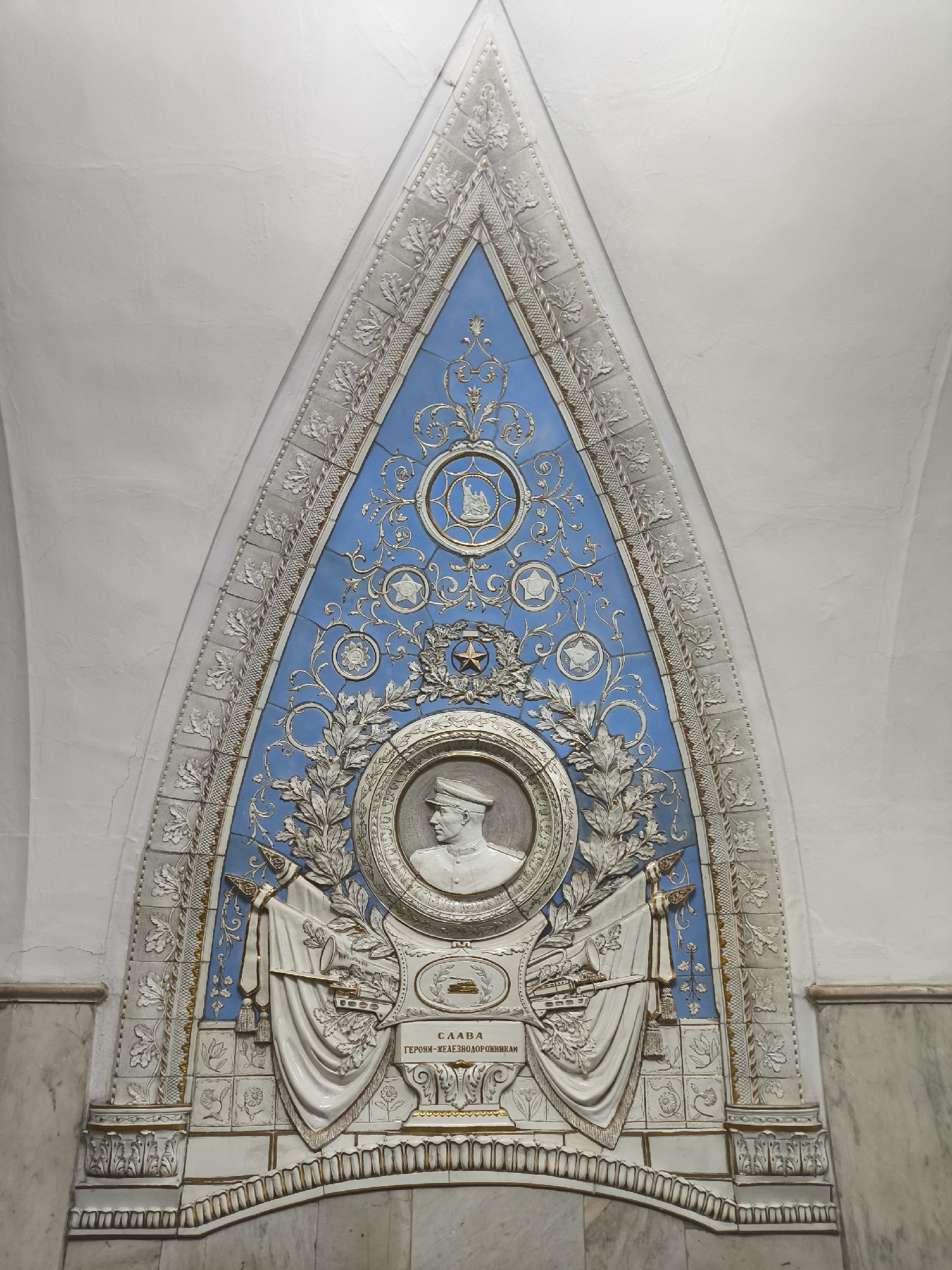 The image captures a decorative panel within a building. The panel is primarily white and blue, arranged in an arched shape. The central element is a circular relief of a person wearing a hat, surrounded by a laurel wreath. Above the portrait, there are decorative patterns, including stars and geometric designs. Below the portrait, a banner drapes across two crossed objects. The banner features writing in a language that cannot be determined. The background of the panel is a light blue color, and the surrounding border is white. The panel appears to be mounted on a wall with a marble base.