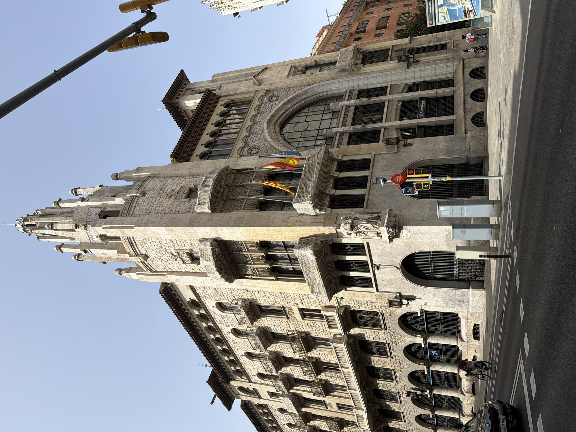 The image shows a large, ornate stone building in Barcelona, Spain, with intricate architectural details. The building features multiple stories with arched windows, balconies with wrought-iron railings, and a prominent tower with a spire. Two flags are visible on the building, one yellow and red striped and another with a red, yellow, and blue design. A "CaixaBank" sign is visible on the building. In the foreground, a street with a pedestrian crossing and traffic lights is present. A dark-colored car is driving on the street, and a person on a bicycle is riding on the sidewalk. Two people are standing on the sidewalk in front of the building. The sky is clear and blue.