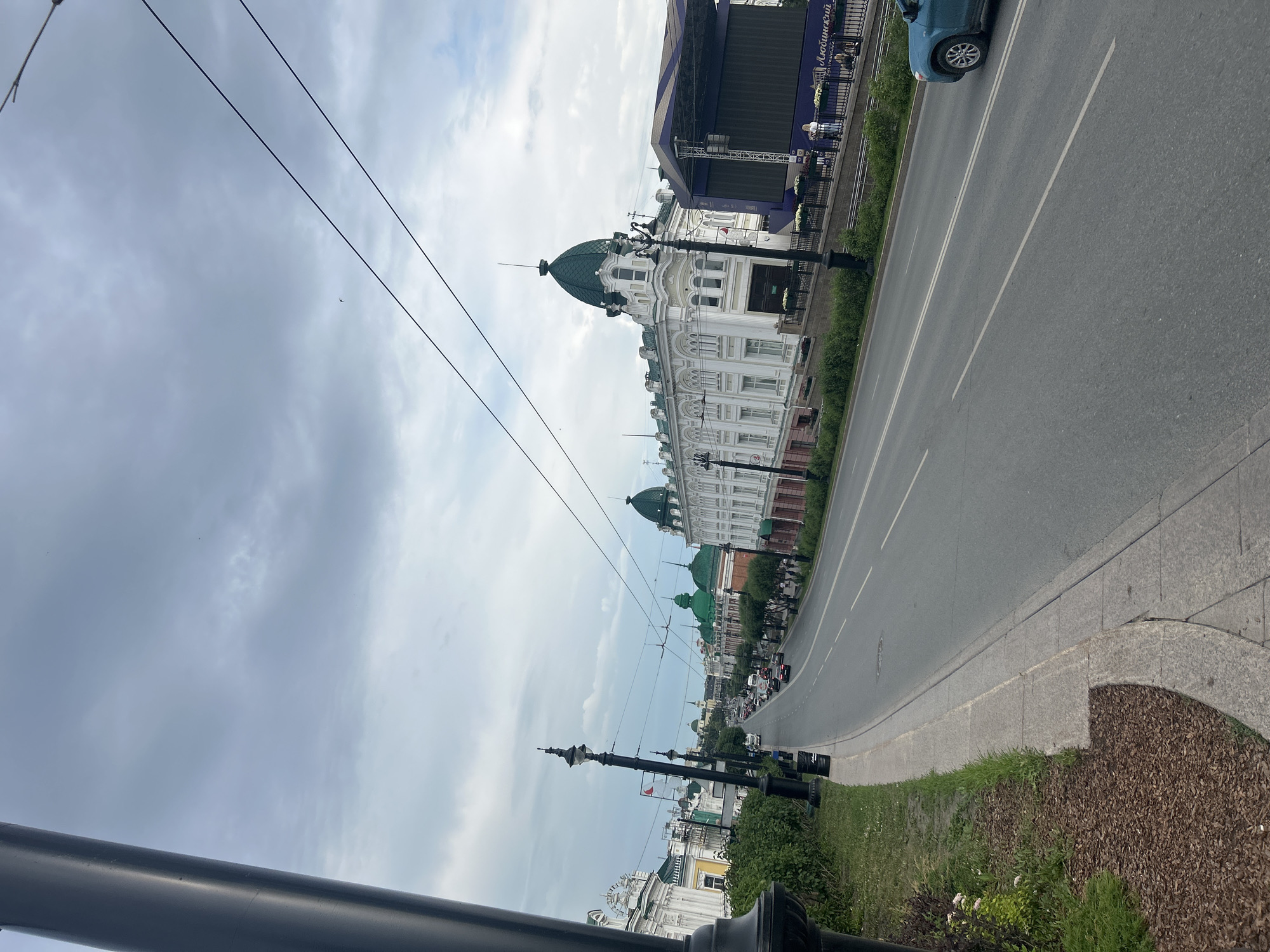 This image shows a street scene in Omsk, Russia, likely on Lyubinsky Prospekt. In the foreground, a dark grey lamppost stands on the left side of the frame, partially obscuring the view. The street curves to the right, with a sidewalk and green space on the left. Several cars are driving on the asphalt road, which has white lane markings.
Along the right side of the street, there are ornate buildings with white facades and green domed roofs. One prominent building features large windows and decorative architectural elements. To the far right, a large dark blue stage with a screen is set up, possibly for an event. Some people are visible near the stage and along the sidewalk. The sky is overcast with grey clouds. The overall impression is an urban environment with historical architecture and signs of modern activity.