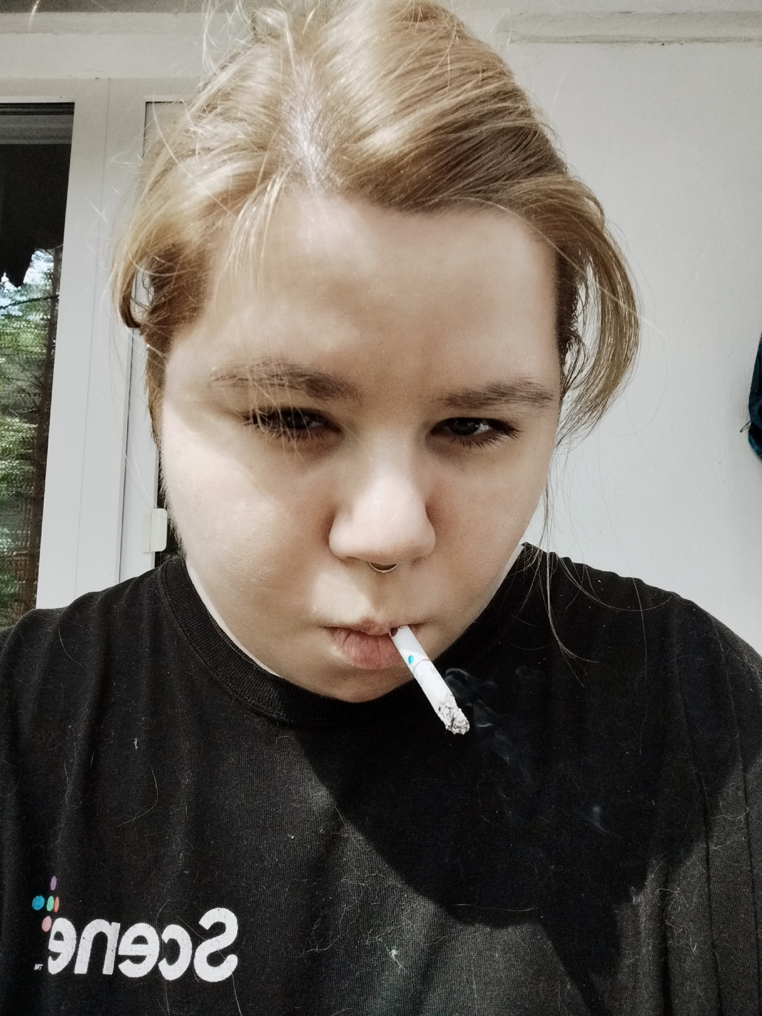 A person with blonde hair pulled back is shown from the chest up, smoking a cigarette. They are wearing a black t-shirt with a white logo that reads "scene" in reverse. The person has a septum ring. The background is out of focus, showing a white wall and a window with greenery visible through it. The lighting suggests an indoor or semi-outdoor setting.