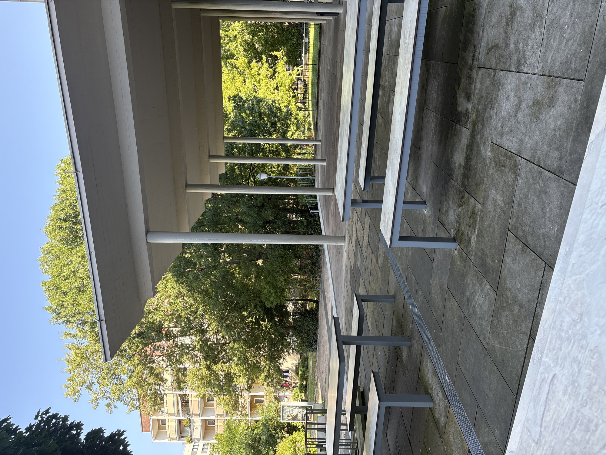 The image shows an outdoor seating area with concrete paving and a covered section supported by columns. Several picnic tables with benches are arranged on the paving. In the background, there are trees, a multi-story apartment building, and a lamppost. The scene appears to be in an urban environment, possibly a park or public space within the city of Dresden, Germany. There are no people visible in the image. The weather appears to be clear and sunny, with a blue sky overhead.
