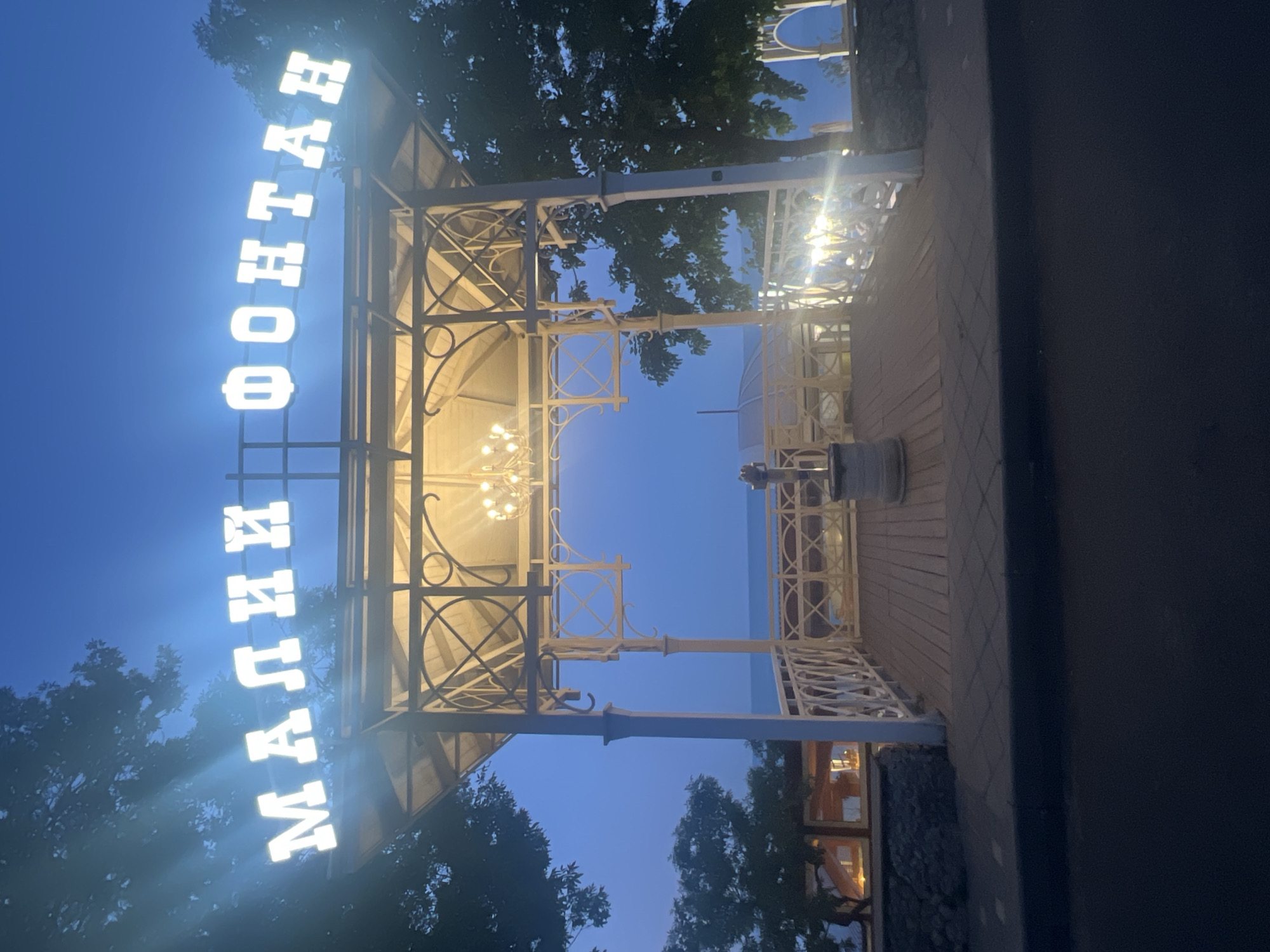The image depicts the illuminated sign "МАЛІЇ ФОНТАН" above a white gazebo at dusk. The gazebo has a wooden floor and a decorative railing. Inside the gazebo, a chandelier is visible. Beyond the gazebo, the Black Sea can be seen. The scene is located on the coast of Odesa, Ukraine. The sky is a deep blue, indicating twilight. There are trees on either side of the gazebo. The lighting from the sign and the gazebo creates a warm glow.