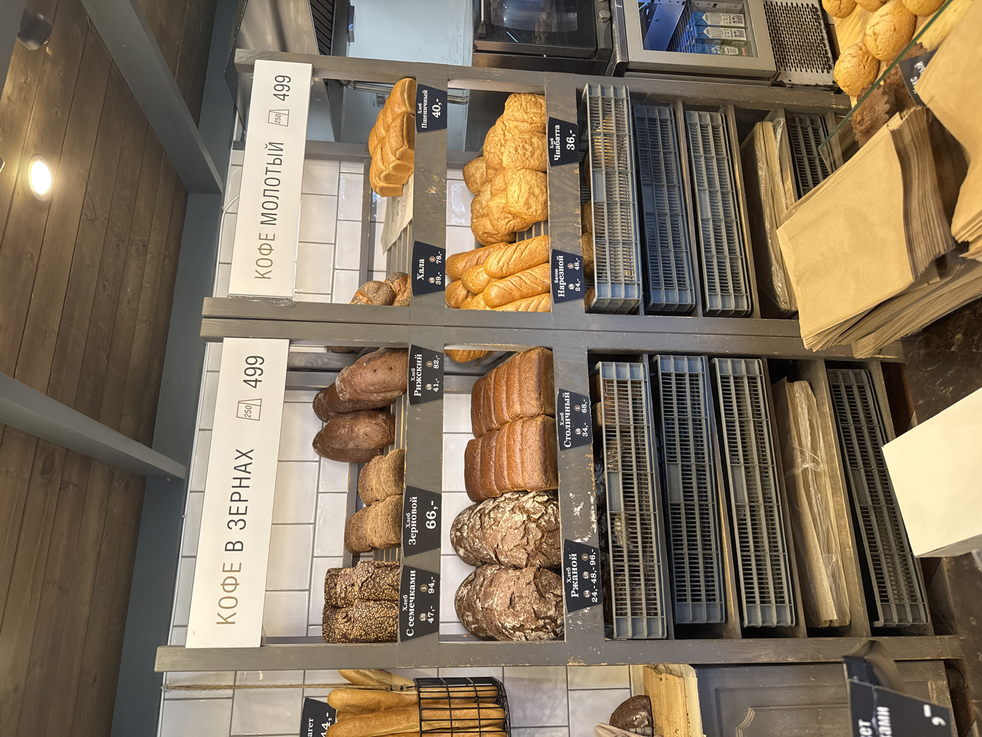The image depicts the interior of a bakery, likely located in Ladozhskoe Ozero, Russia, as indicated by the Russian text on the signage. The focus is on a display of baked goods and coffee. On the left side, a sign reads "КОФЕ В ЗЕРНАХ" (Coffee Beans) with a price of 499. Below this are shelves filled with various types of bread, each with labels and prices in Russian. Examples include "Хлеб С семечками" (Bread with seeds) priced at 47, 94, and "Хлеб Зерновой" (Grain Bread) for 66. The right side displays a sign for "КОФЕ МОЛОТЫЙ" (Ground Coffee) also priced at 499. This section also features loaves of bread and rolls. Items visible include "Хлеб Пшеничный" (Wheat Bread) for 40, "Хала" (Challah) for 39, 78, and "Хлеб Чиабатта" (Ciabatta Bread) for 36. In the foreground, stacks of brown paper bags are arranged, suggesting they are for customer use. The overall environment appears clean and organized, typical of a retail food establishment. The lighting is bright, illuminating the products.