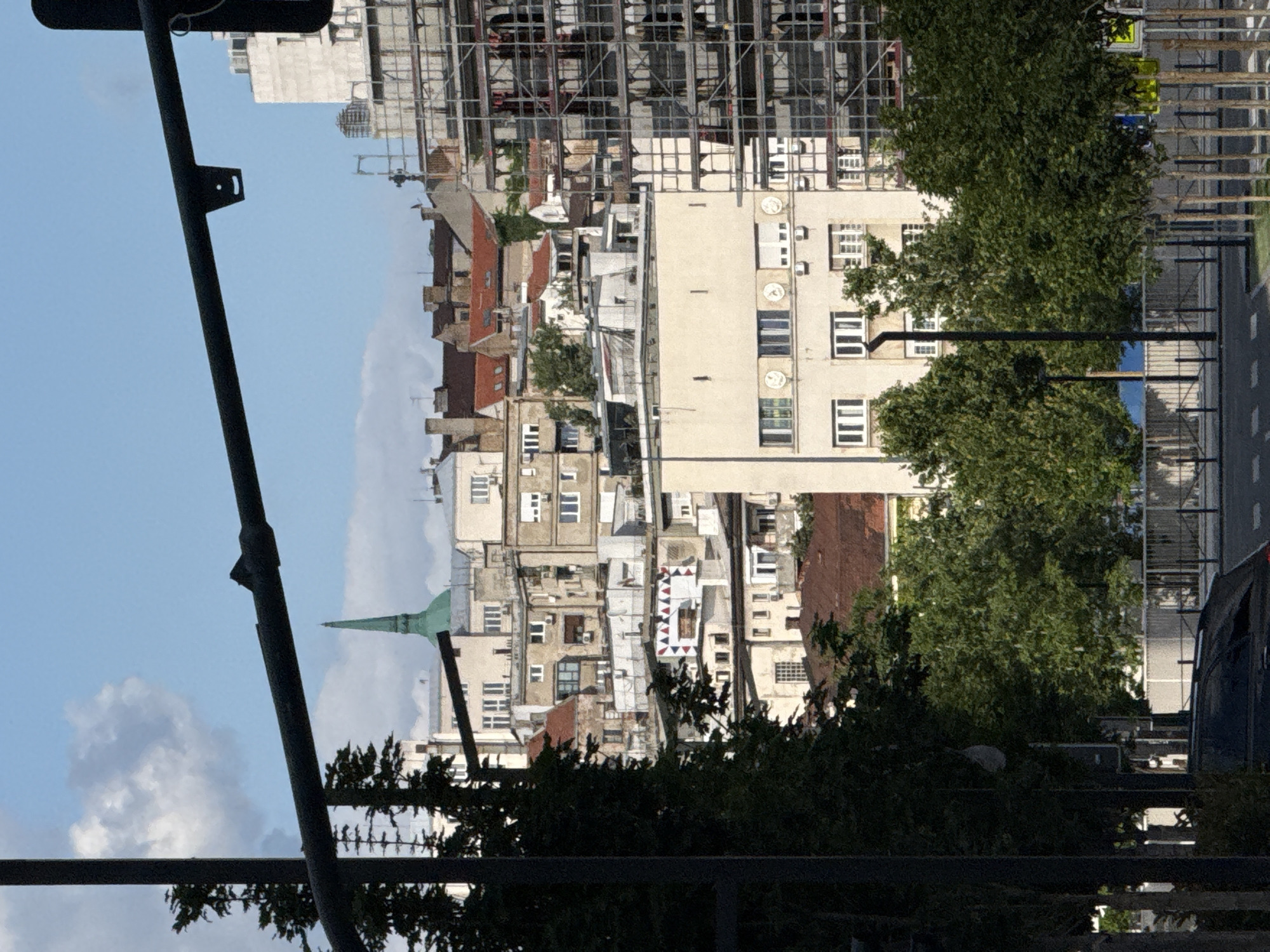 This is a photograph of a cityscape in Belgrade, Serbia, likely taken from a street level looking upwards. The scene is dominated by a dense collection of multi-story buildings, some appearing to be under construction or renovation, evidenced by extensive scaffolding on the right side of the frame. The architecture varies, with some buildings featuring older, traditional styles with red-tiled roofs, while others are more modern, with lighter colored facades and numerous windows. A prominent green spire, possibly belonging to a church or historical building, is visible in the background, cutting through the skyline against a clear blue sky with scattered white clouds. Lush green trees line the foreground and mid-ground, obscuring parts of the lower levels of the buildings and creating a natural contrast with the urban environment. A dark-colored car is partially visible in the lower left corner, suggesting a street or parking area. Traffic lights and street lamps are also present, indicating a functioning urban thoroughfare. The overall impression is of a bustling city with a mix of old and new architectural elements, set against a bright, sunny day.