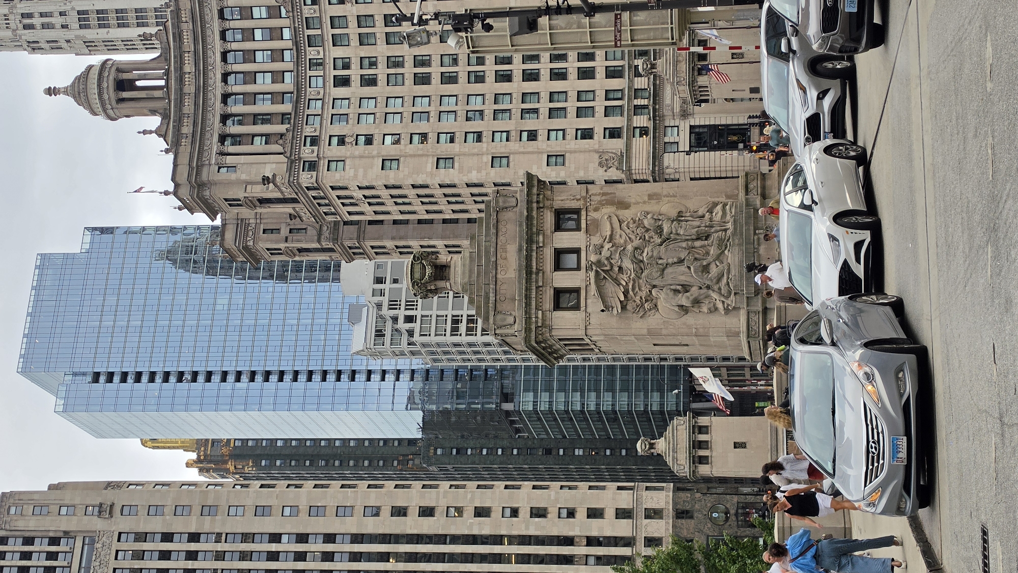 The image depicts a street view in Chicago, Illinois, USA, featuring a confluence of architectural styles and urban activity. Dominating the background are several high-rise buildings, including a modern glass skyscraper and older, ornate stone structures. In the foreground, a stone monument with a bas-relief sculpture and the inscription "CITY OF CHICAGO" is prominently displayed. Several cars, including a silver Hyundai sedan and a white sedan, are on the street, suggesting traffic. Pedestrians are visible on the sidewalks and near the monument, some appearing to be tourists or locals going about their day. The scene is captured on an overcast day, with the sky a muted grey. The overall impression is one of a bustling urban environment where historical architecture meets contemporary design.