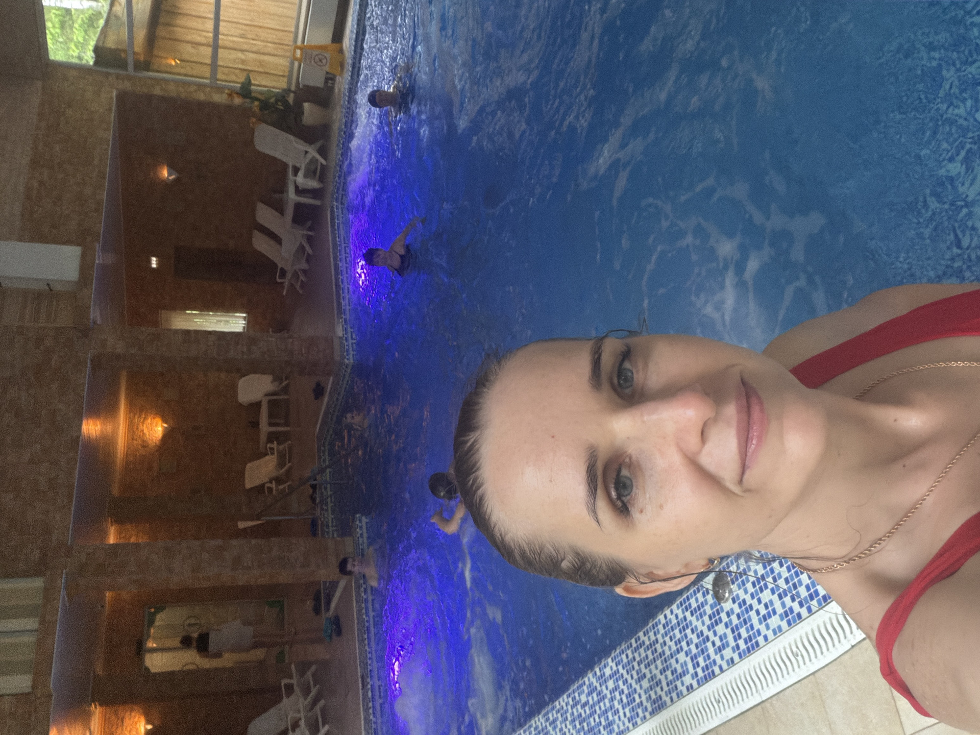 A woman is taking a selfie in what appears to be an indoor swimming pool. She is wearing a red swimsuit and a gold necklace. The pool has blue tiled walls and is lit with blue underwater lights. There are other people swimming in the pool in the background. There is a seating area with lounge chairs visible along the side of the pool. The location is Zaplav'e, Russia.
