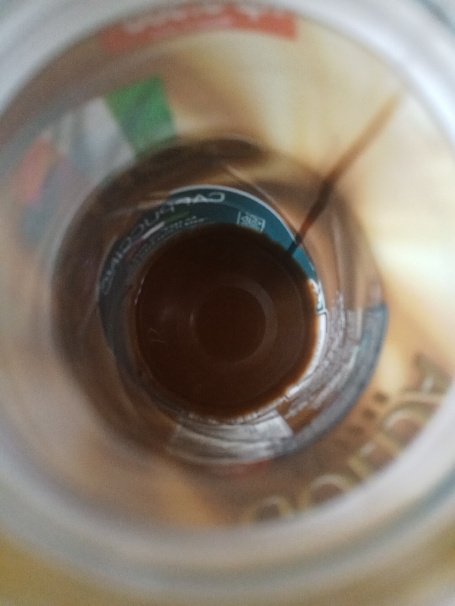 The image shows the inside of a glass jar with some dark liquid at the bottom. The jar has some labels visible, one of which appears to say "CAPPUCCINO". The background is blurry and shows hints of other objects and colors, possibly suggesting an indoor environment. The scene does not depict any specific location in the city of Buketglumpang, Indonesia, as there are no discernible landmarks or contextual clues to support such a claim.